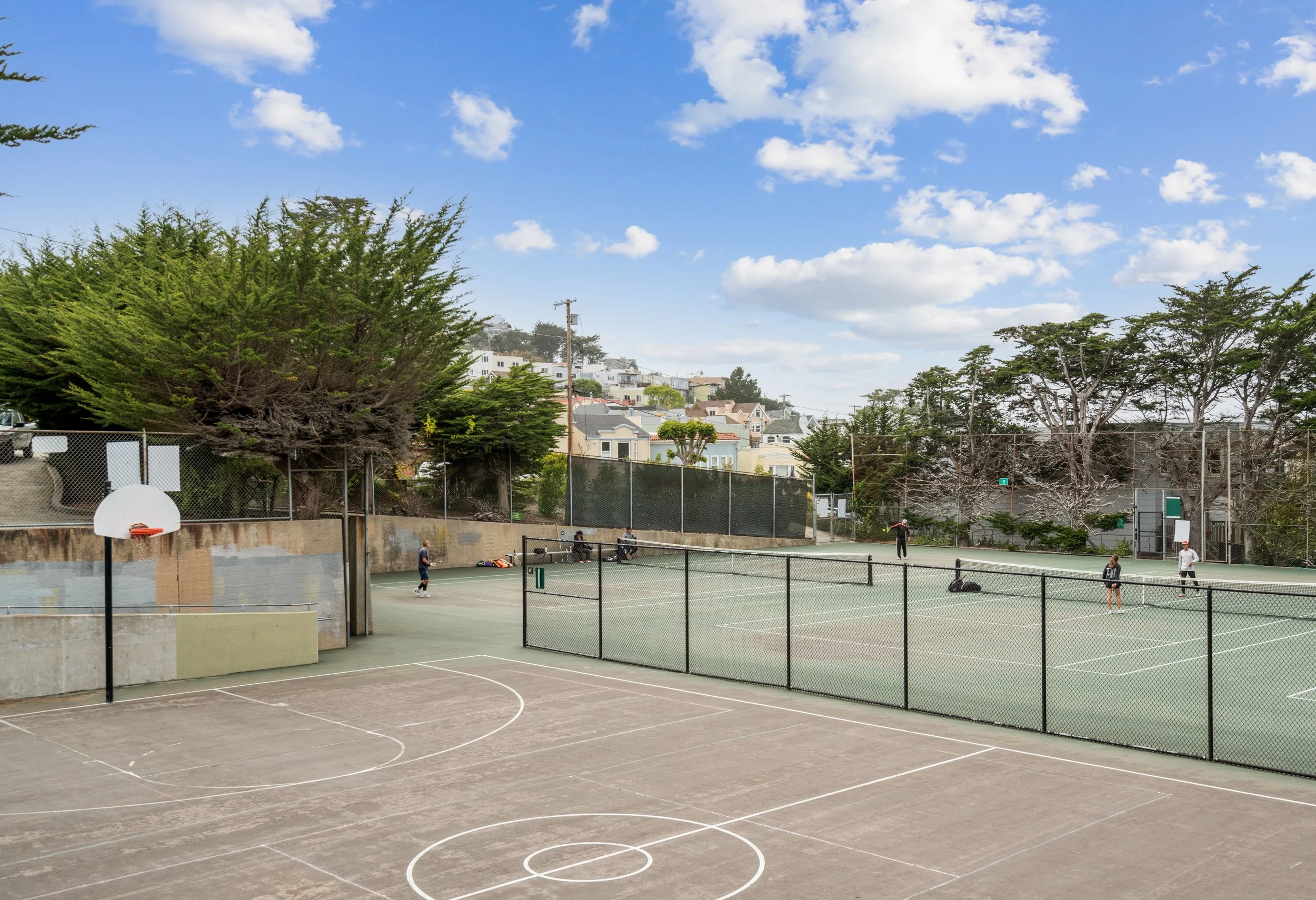 An outdoor tennis court with a chain-link fence, surrounded by trees and residential buildings under a partly cloudy sky.