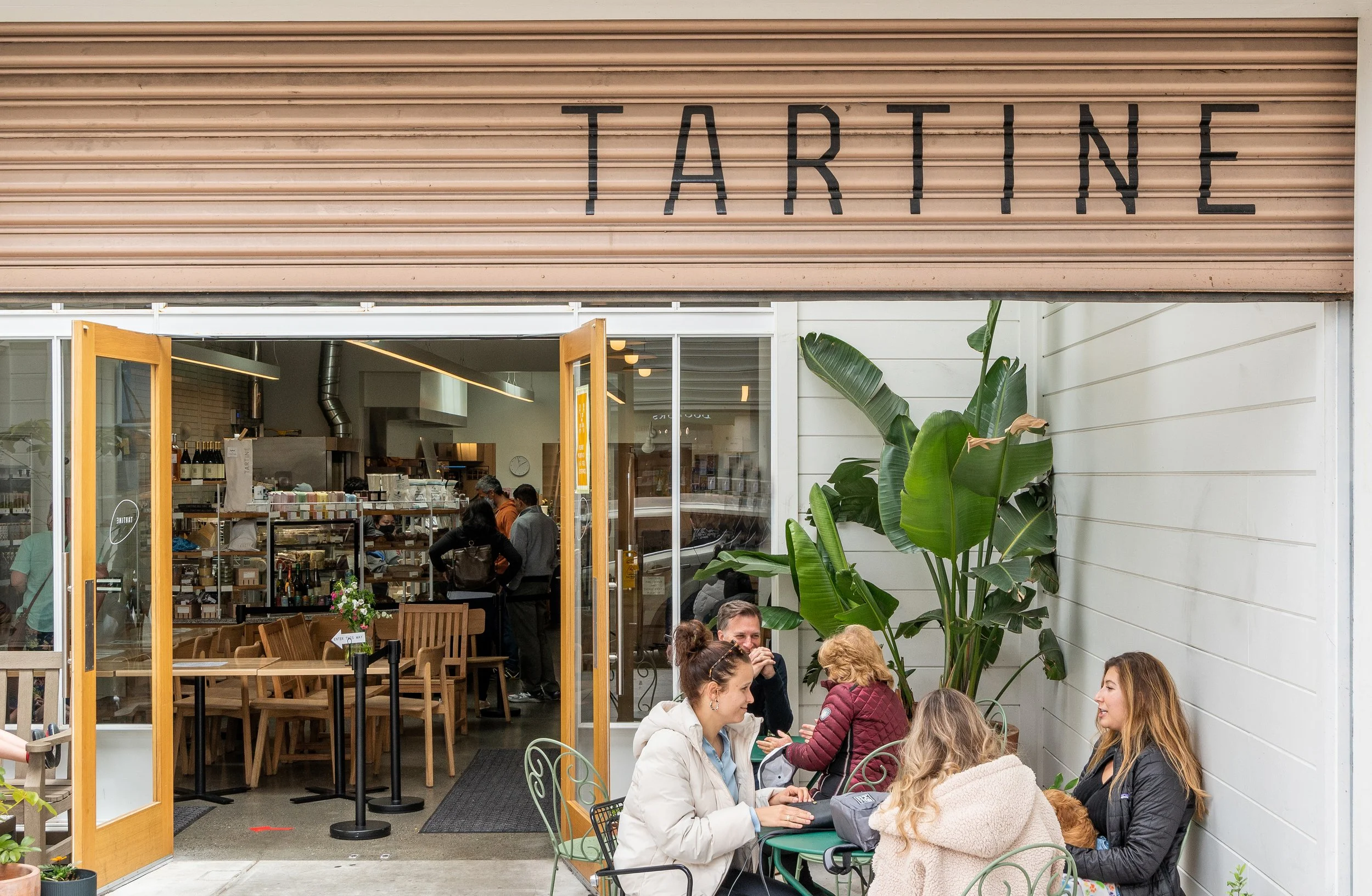 The exterior of a café with an open entrance, outdoor seating area with people sitting and chatting, large green plants, and inside visible kitchen and dining areas.
