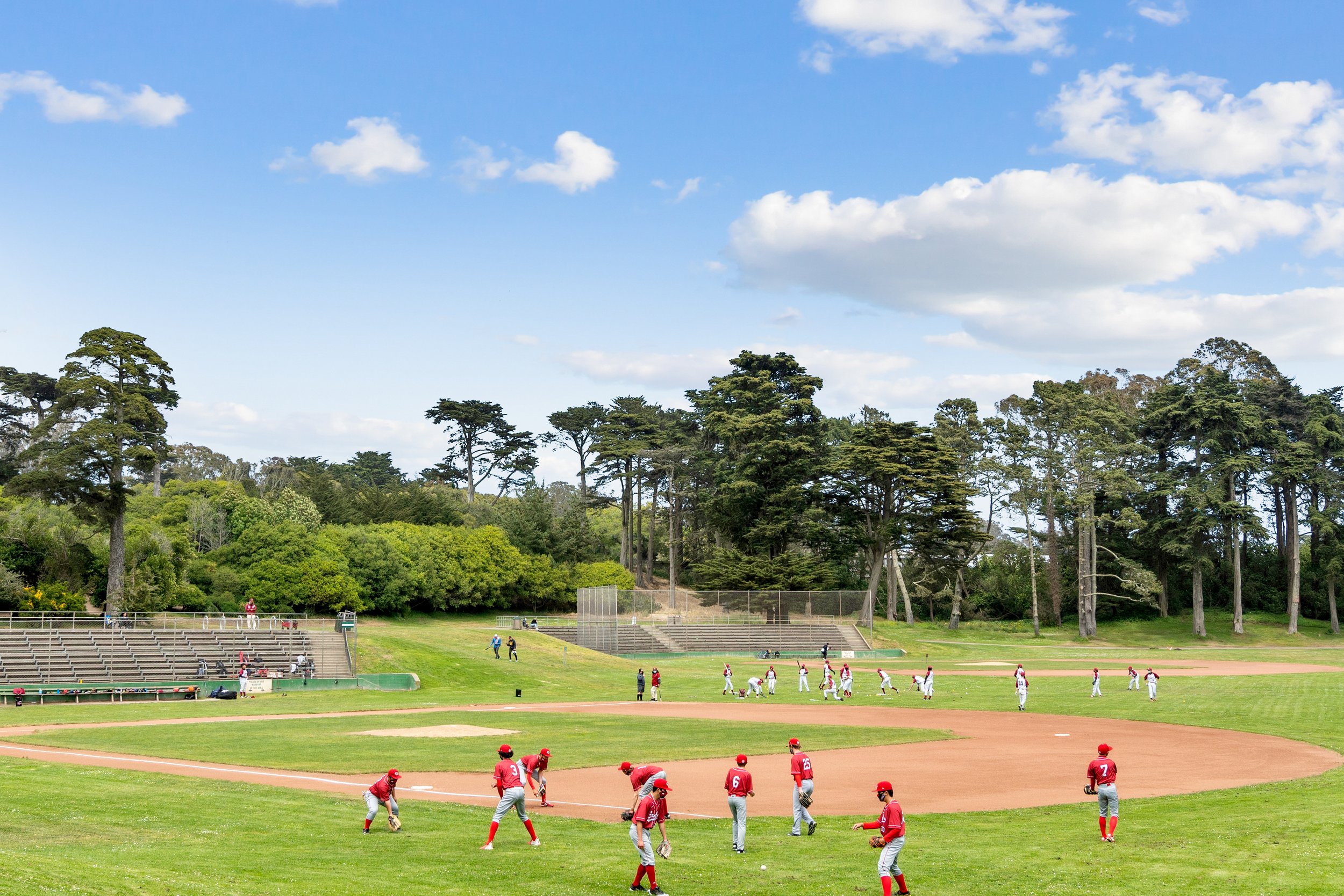 A baseball game taking place at a park with players warming up on the field, surrounded by trees and blue sky with clouds.