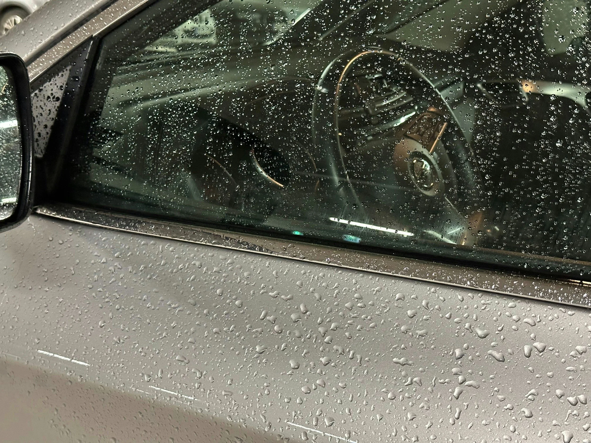The image shows the front windshield of a car covered with water droplets at a car wash, with a view of the car's clean interior including the steering wheel and dashboard.
