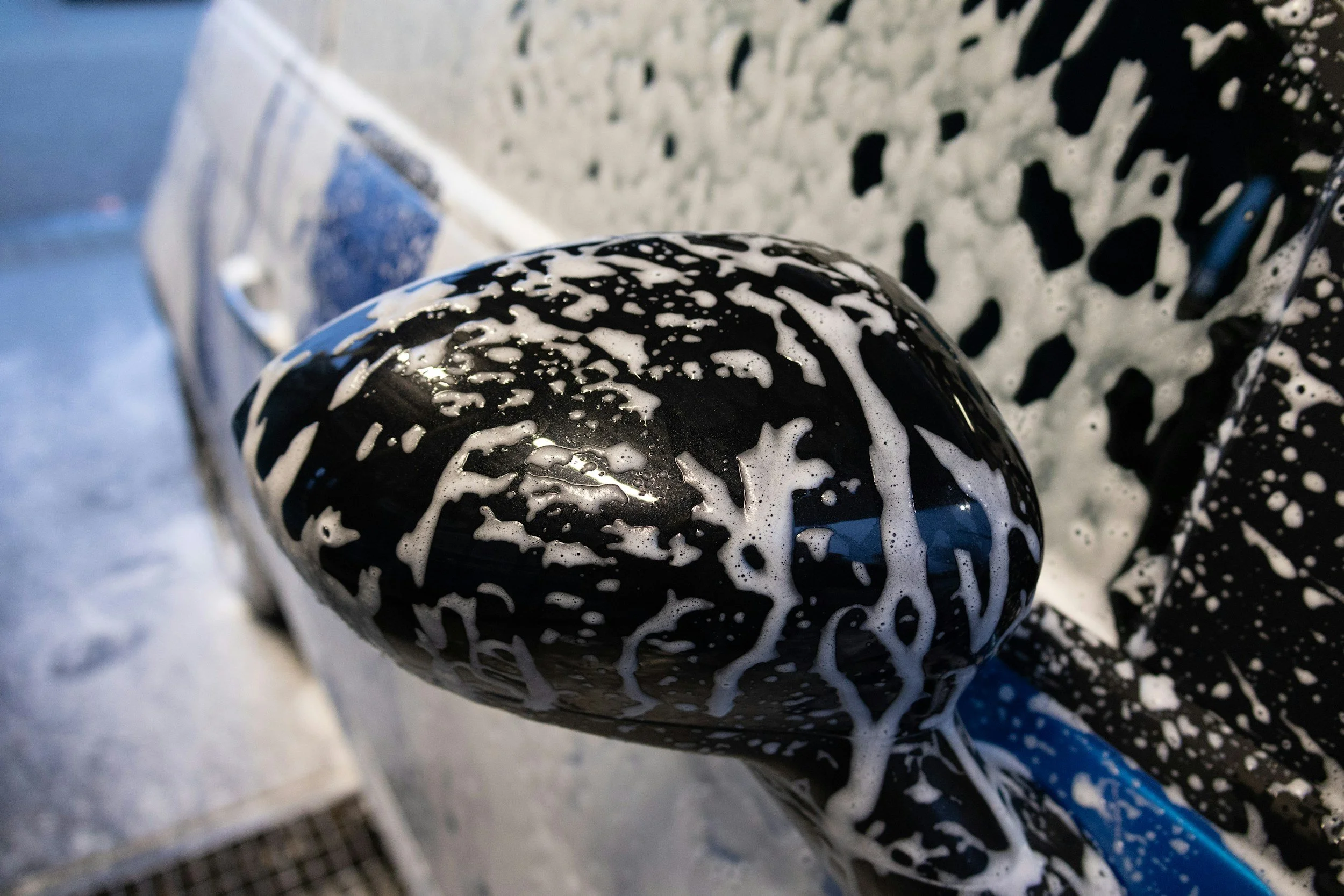 Close-up of a black object, possibly a washing machine or appliance, with white soap or foam on its surface. There is a silver-colored item and a blue water hose visible in the background.