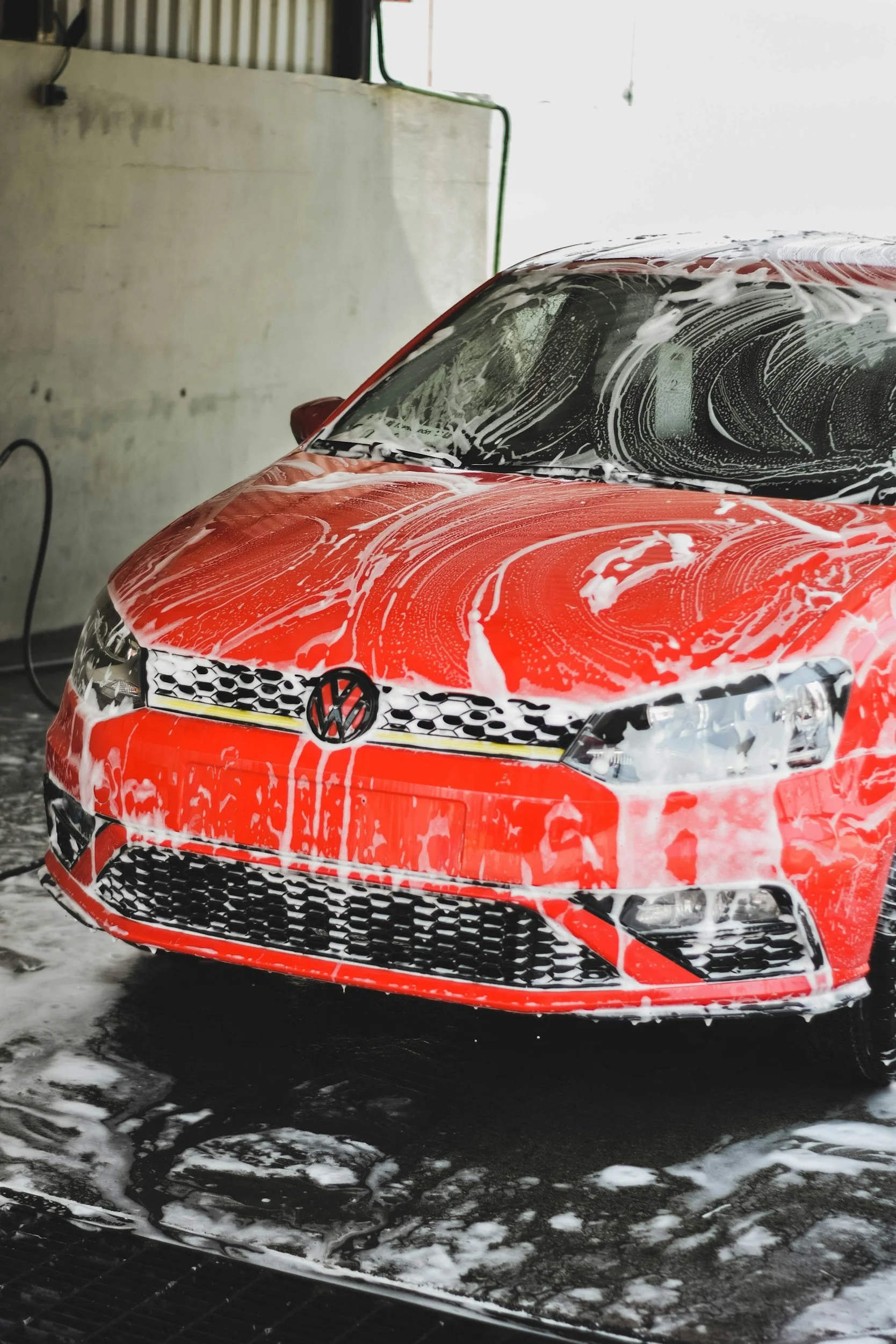 A red Volkswagen car being washed with soap, foam, and water in an indoor car wash station.