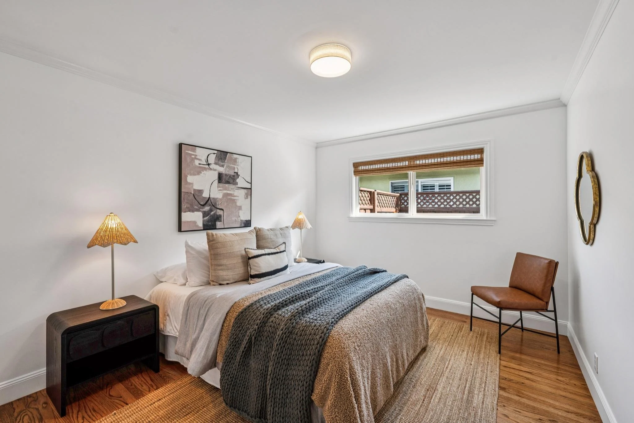 A bedroom with a bed, pillow, throw blanket, two lamps, a framed abstract art piece, a window with bamboo blinds, and a brown chair against the wall.