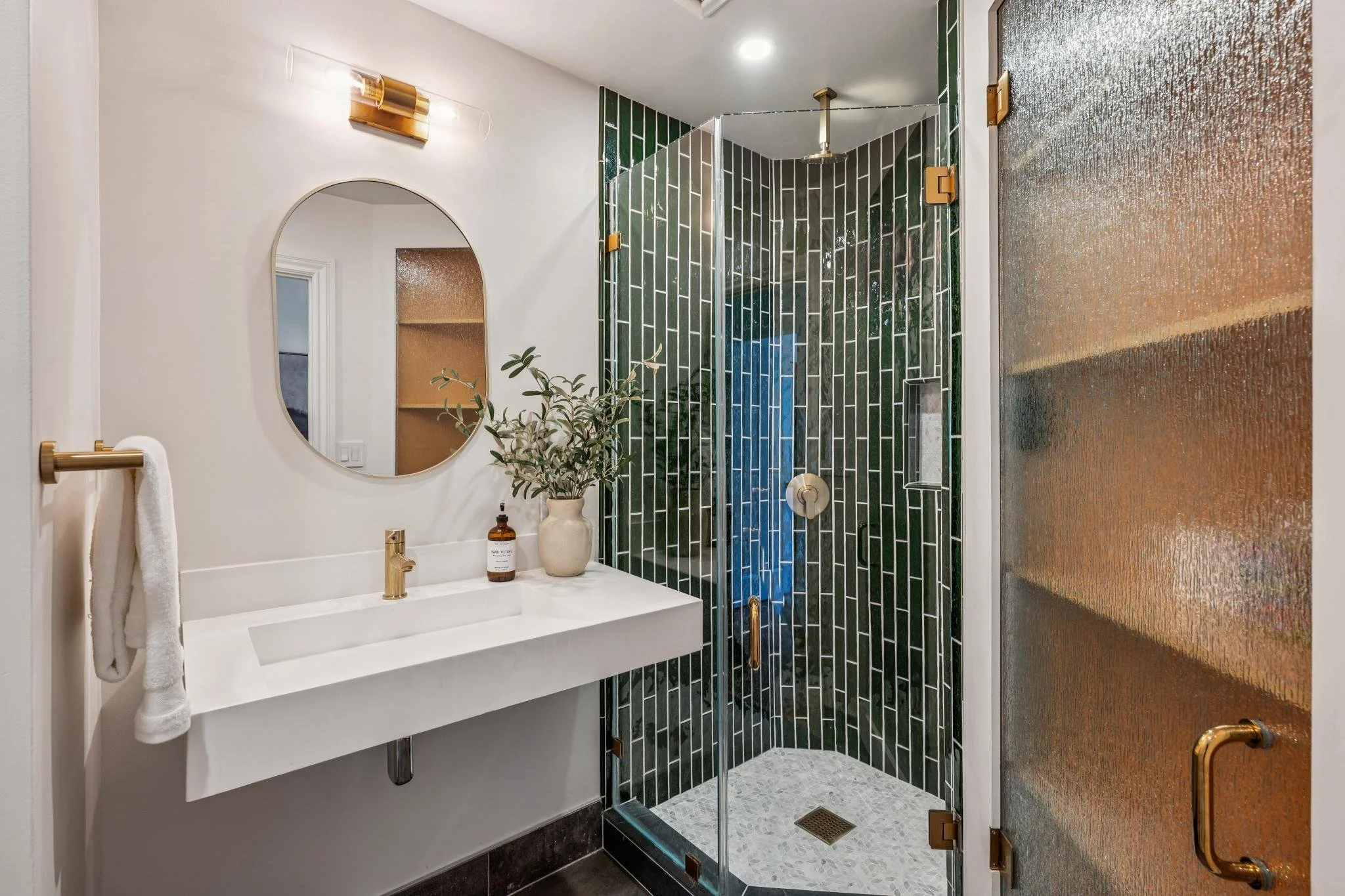 A modern bathroom featuring a white vanity with a round mirror, a green tiled walk-in shower with glass doors, a potted plant, and brass fixtures. The bathroom has white walls, dark flooring, and a copper-toned piece of art on the wall.