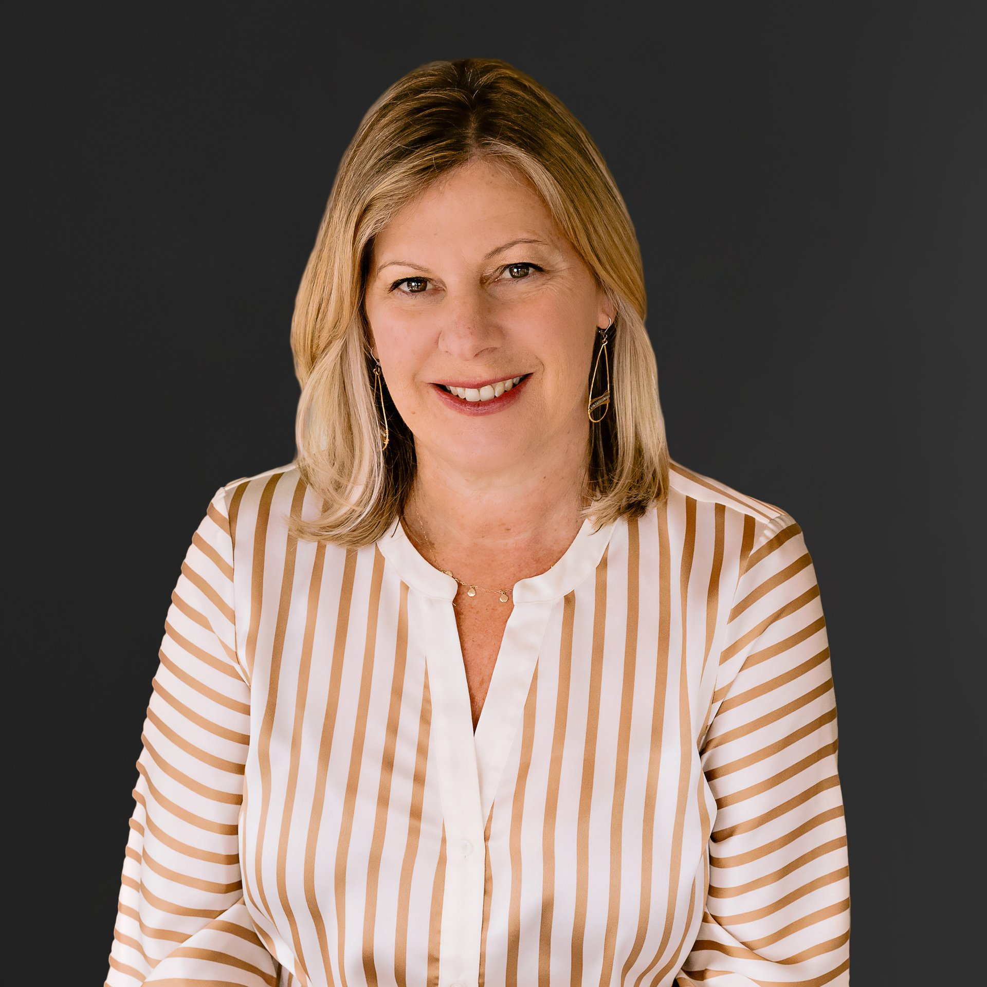 A middle-aged woman with blonde hair and light skin, smiling in front of a dark background, wearing a white and beige striped blouse and gold earrings.