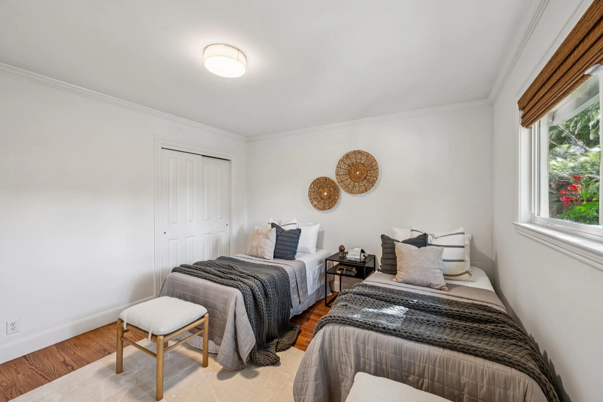 A cozy bedroom with white walls, hardwood floors, and a window with wooden blinds. There are two beds with gray and white bedding, decorative pillows, and a black side table between them. Woven wall decor hangs on the wall above the beds.