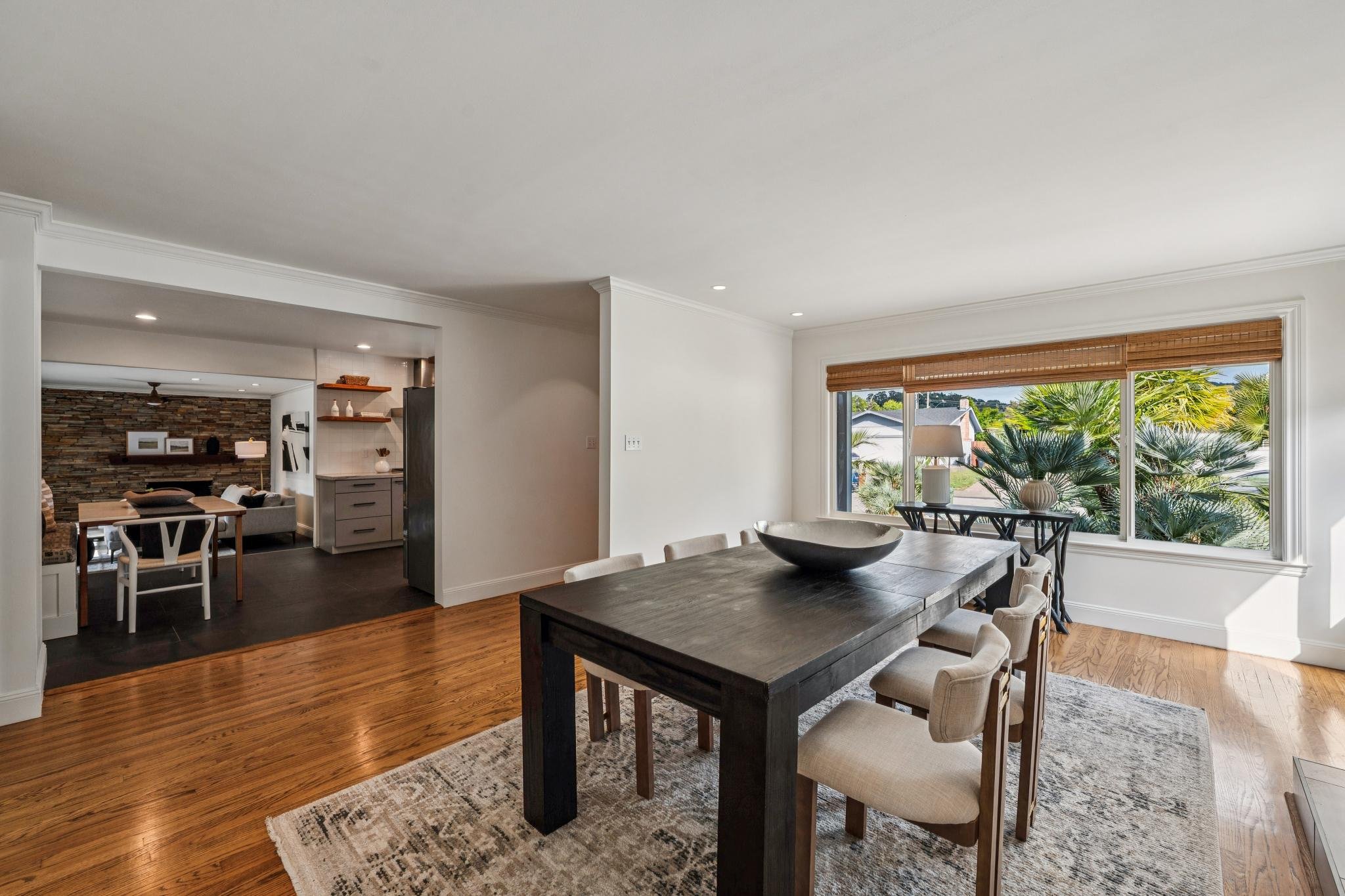 Interior view of a dining room with a wooden table, beige chairs, large window with bamboo blinds, and a view of greenery outside. Adjacent to the dining area is a living space with a stone accent wall, dark wood flooring, and furniture.
