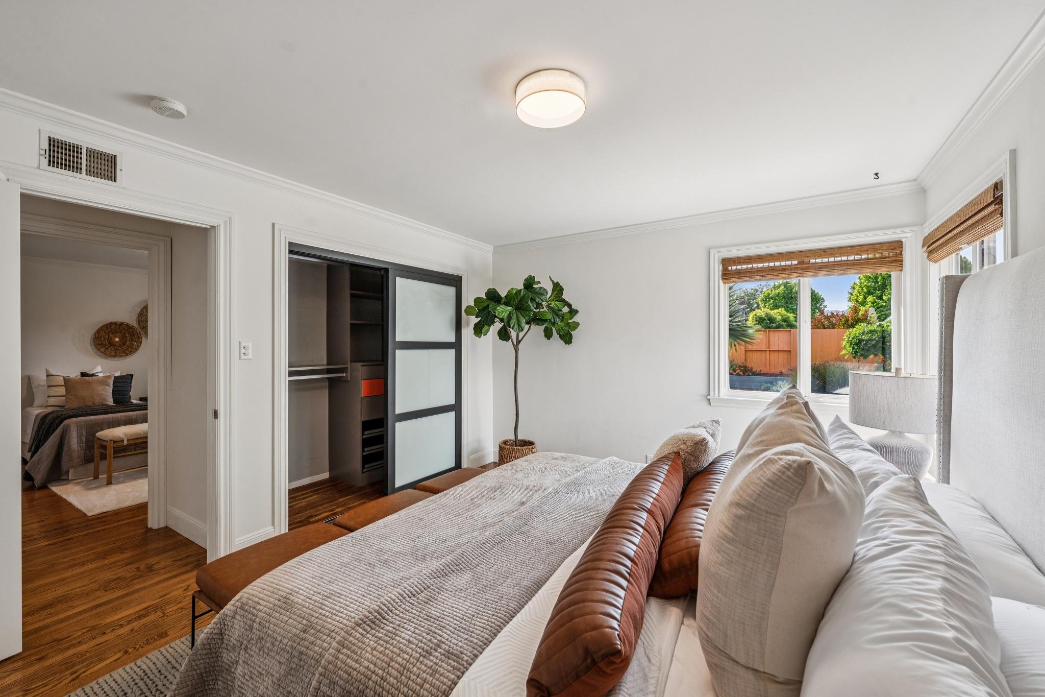 Living room with white walls, a large window with bamboo shades, a beige sofa with white and brown cushions, a potted plant, and a view of a backyard with trees and a wooden fence.