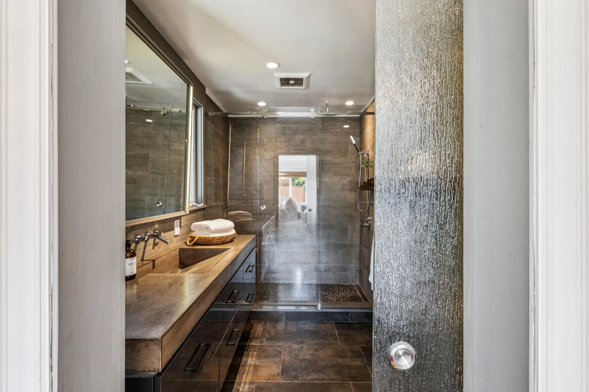 Modern bathroom with a large mirror, wooden vanity, and walk-in shower with glass door, gray tiles, and a rain showerhead.