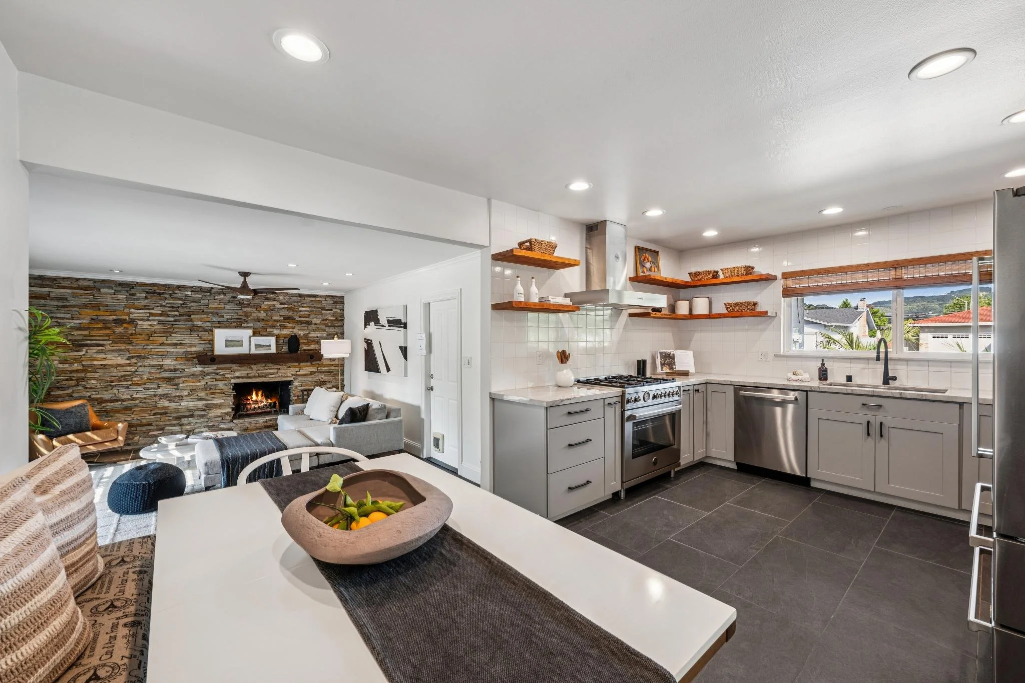 Open-concept kitchen and living room with a stone fireplace, modern appliances, and open shelves with decorative baskets and plates.