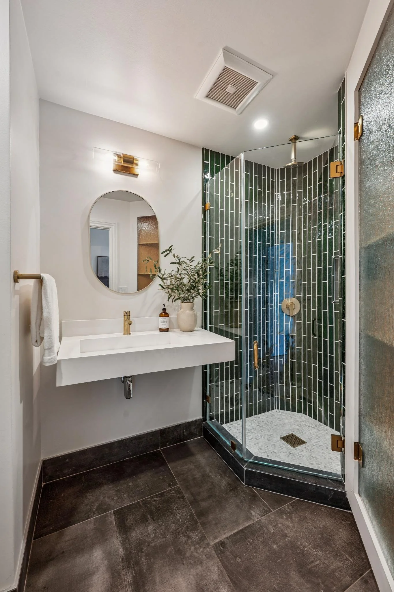 Modern bathroom with a glass shower enclosure, white vanity with a round mirror, dark floor tiles, and a plant on the sink