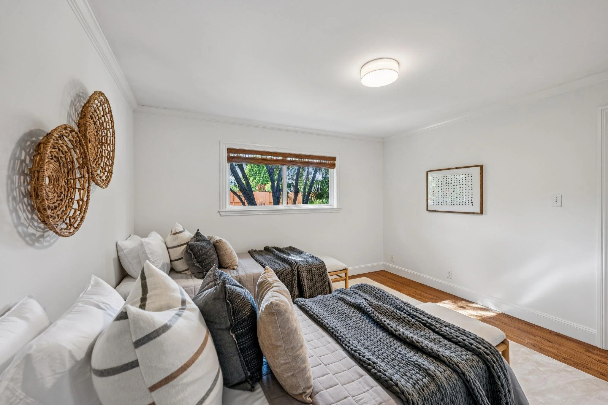 A cozy living room with white walls and hardwood floors. There is a large sofa covered with pillows, a knitted blanket, and a woven bench. A window shows trees outside, and there's a framed picture on one wall and woven wall art on another ceiling wa