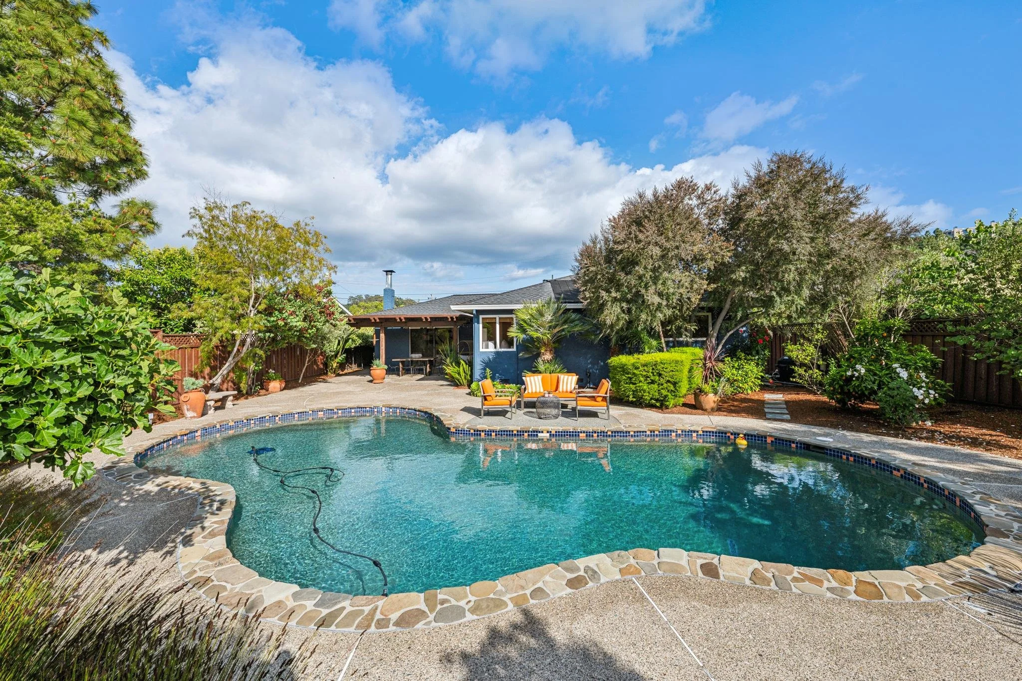 Backyard with swimming pool, patio furniture, trees, and a house under a partly cloudy sky.