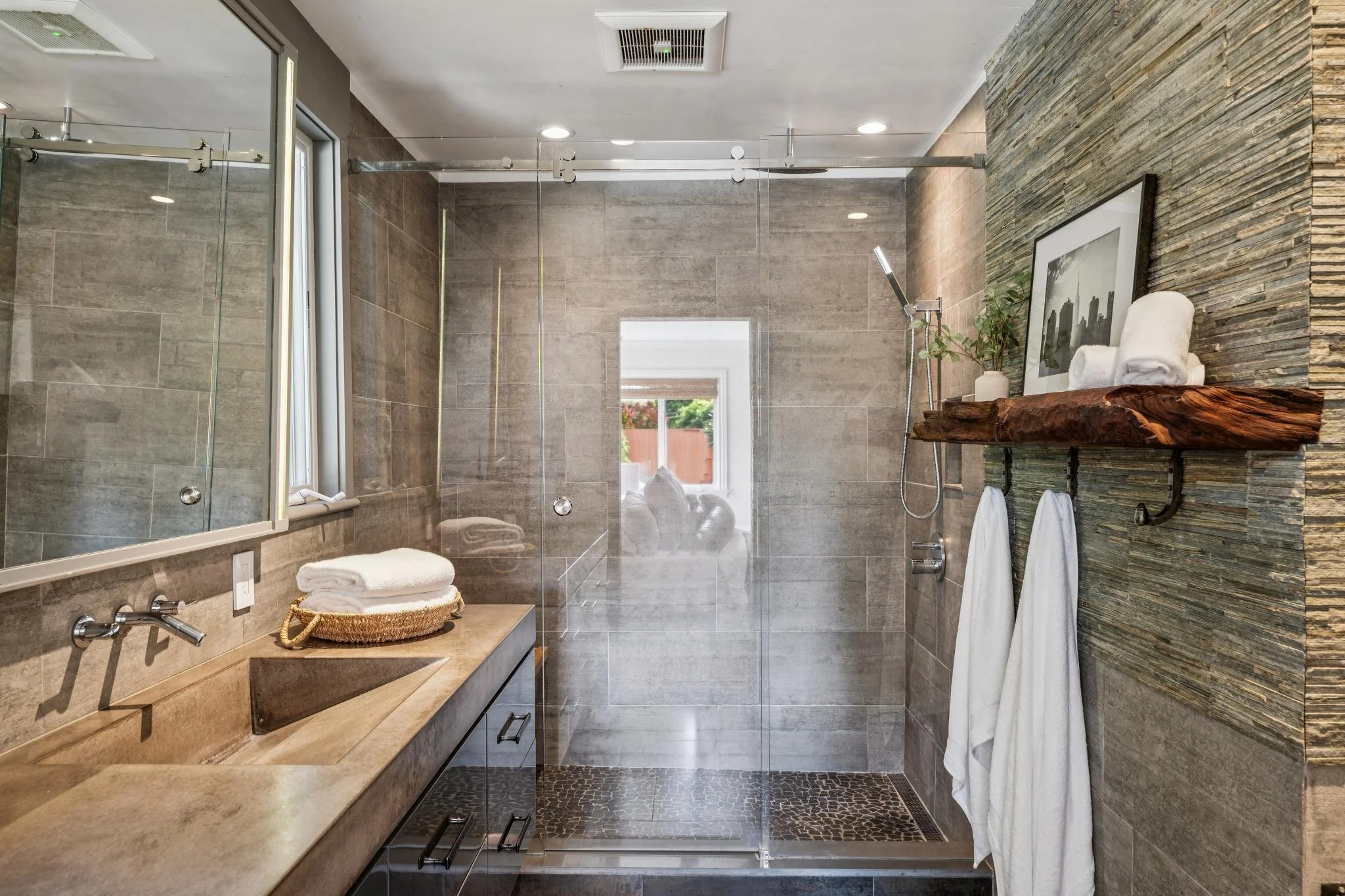 Modern bathroom with beige and gray tiles, glass shower, wooden shelves, and black and white artwork.