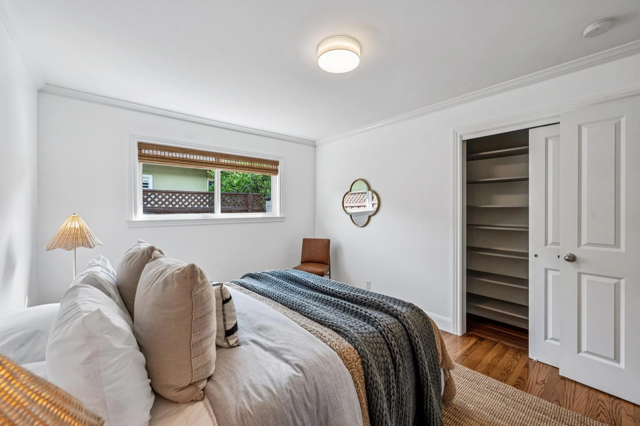 A bedroom with white walls and ceiling, a window with a wooden blind, a bed with beige and white pillows, a black and white blanket, a brown chair, wall mirror, open closet with shelves, and a smoke detector on the ceiling.