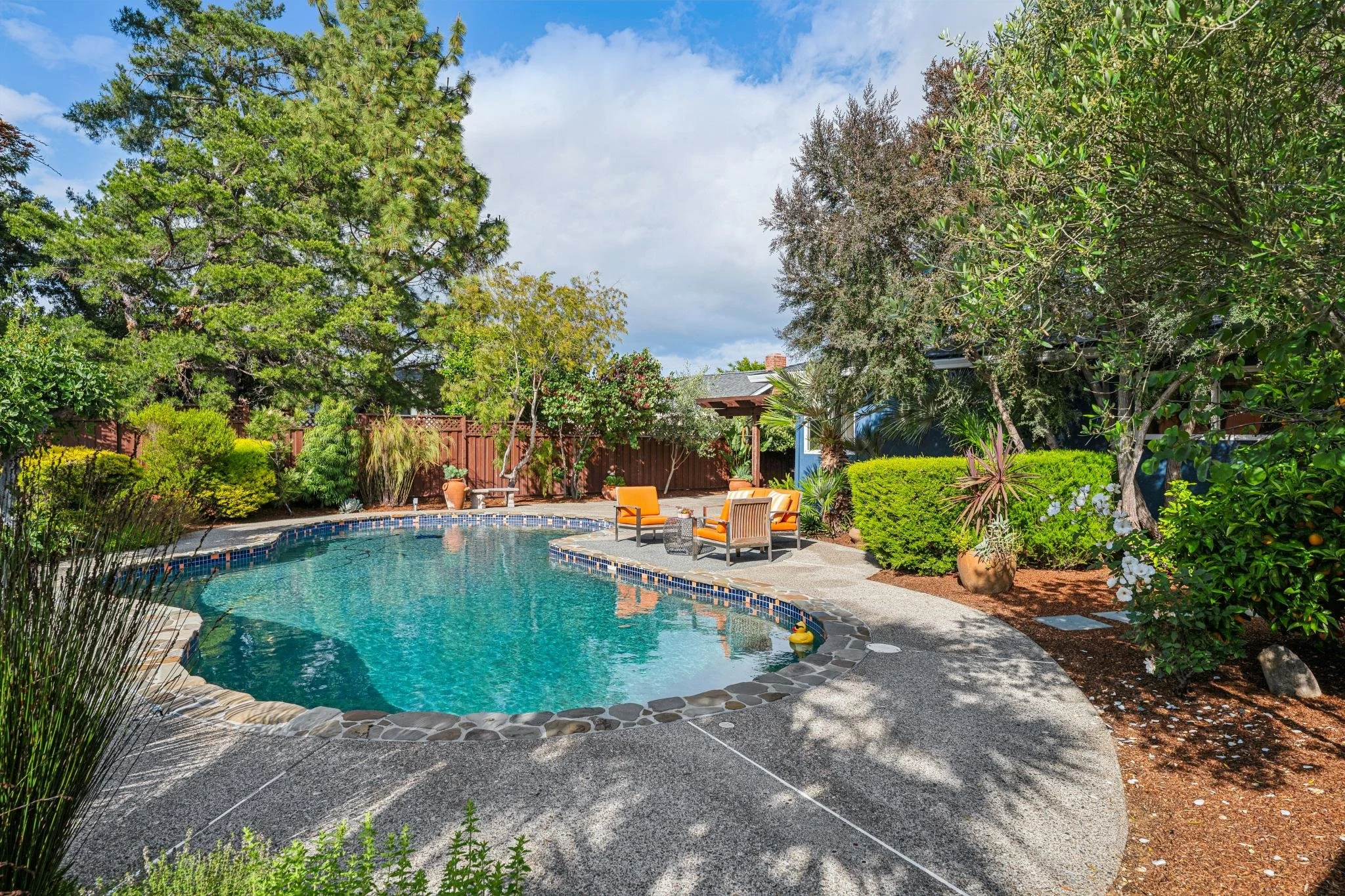 Backyard with a swimming pool surrounded by green trees and bushes, outdoor furniture, and a wooden fence under a partly cloudy sky.