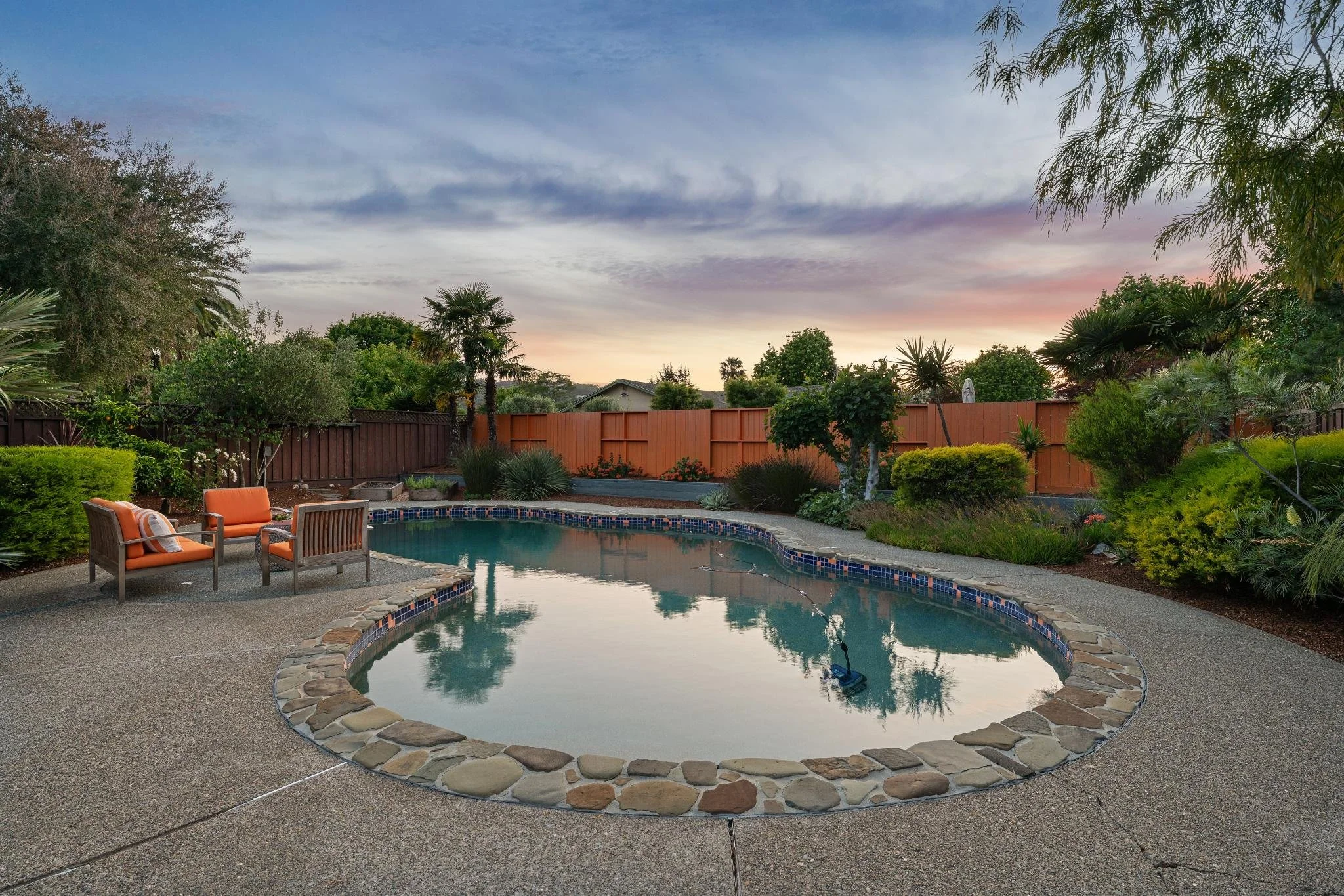 Backyard with a swimming pool, surrounded by chairs, trees, bushes, and a wooden fence, under a colorful sunset sky.