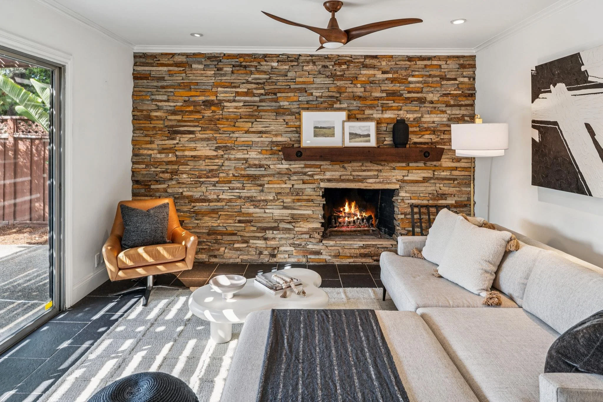 Living room with a fireplace, textured wood wall, and modern decor. There is a white sofa with pillows, an orange armchair, a white coffee table with books, and a sliding glass door revealing outdoor greenery.