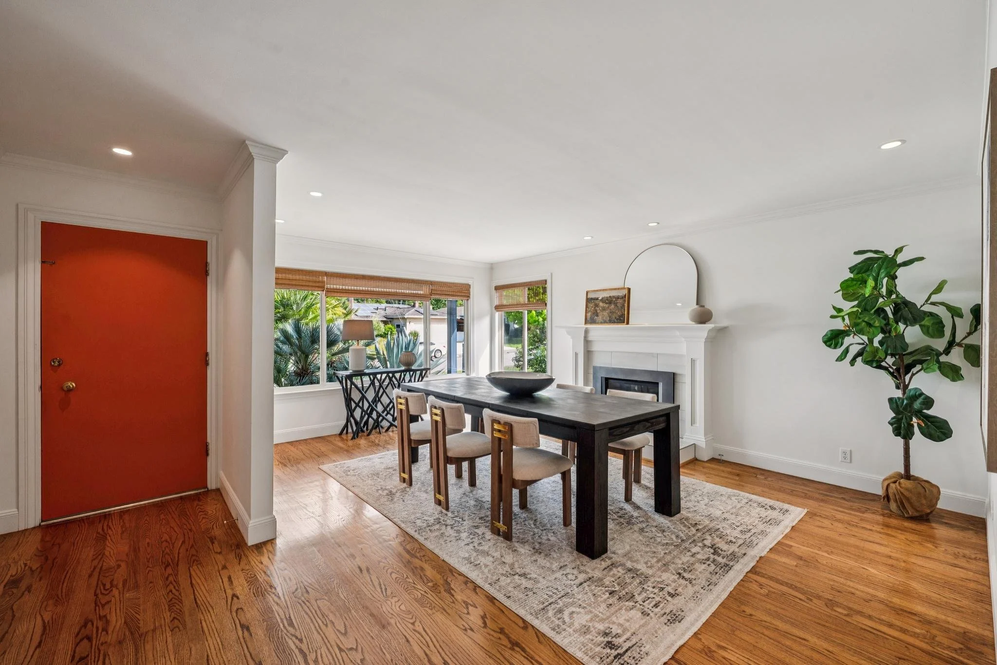 Living room with hardwood flooring, white walls, large windows with blinds, a dark wooden dining table with chairs, a fireplace, plant, and decor on the mantel.