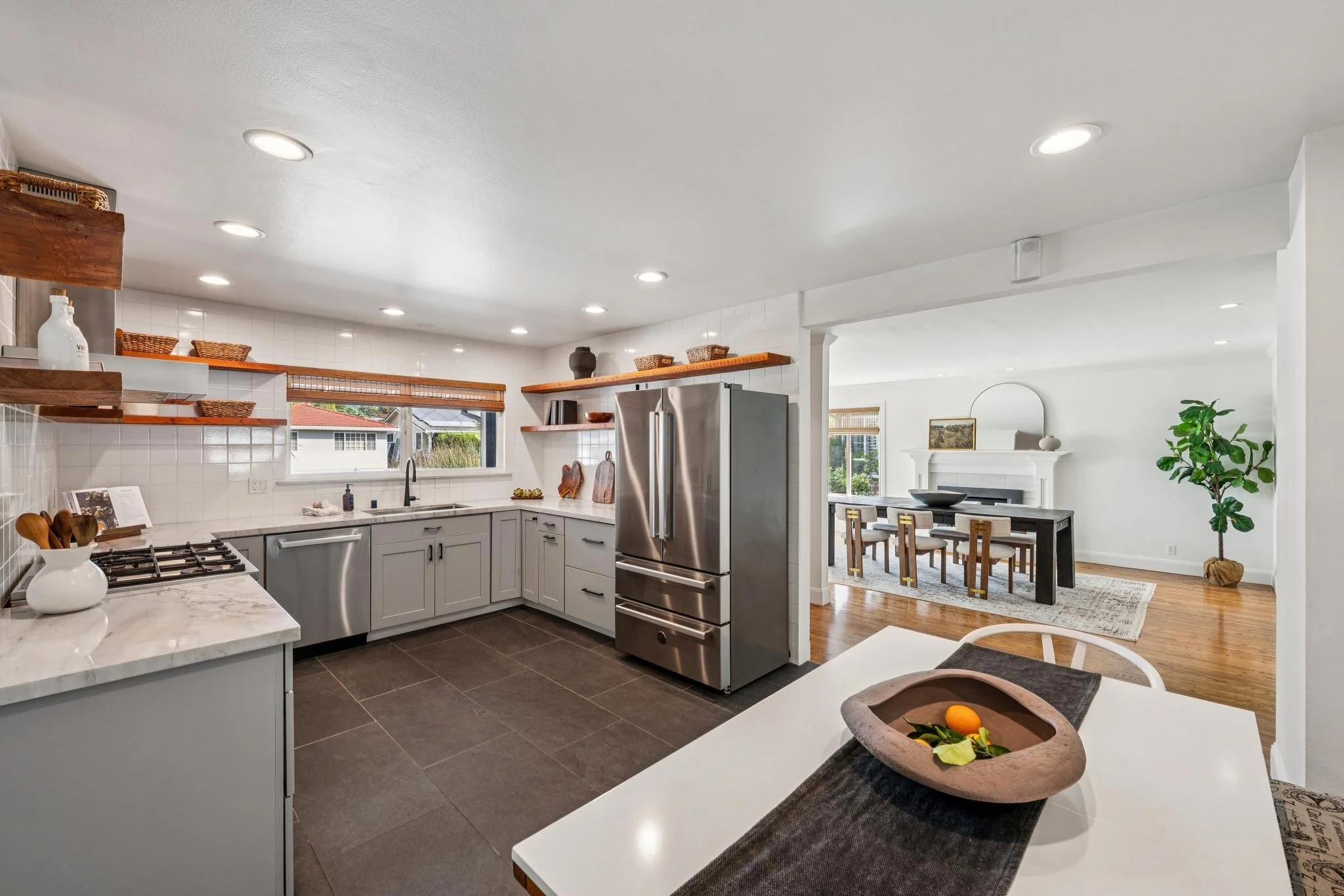 Modern kitchen with white cabinets, stainless steel refrigerator, black countertops, open shelving, and a view into the dining area with a black dining table and chairs. There is a potted plant near a white fireplace in the background.