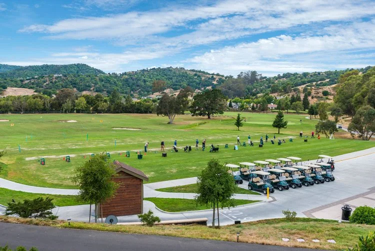 Golf driving range with people practicing swings, lined with carts, and surrounded by trees and rolling hills under a partly cloudy sky.