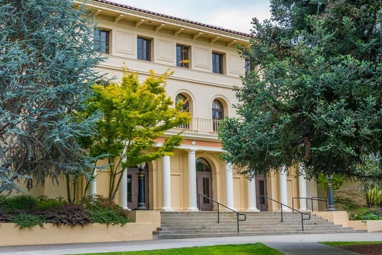 Exterior of a large, historic-style building with tall white columns and a beige facade, surrounded by trees and greenery.
