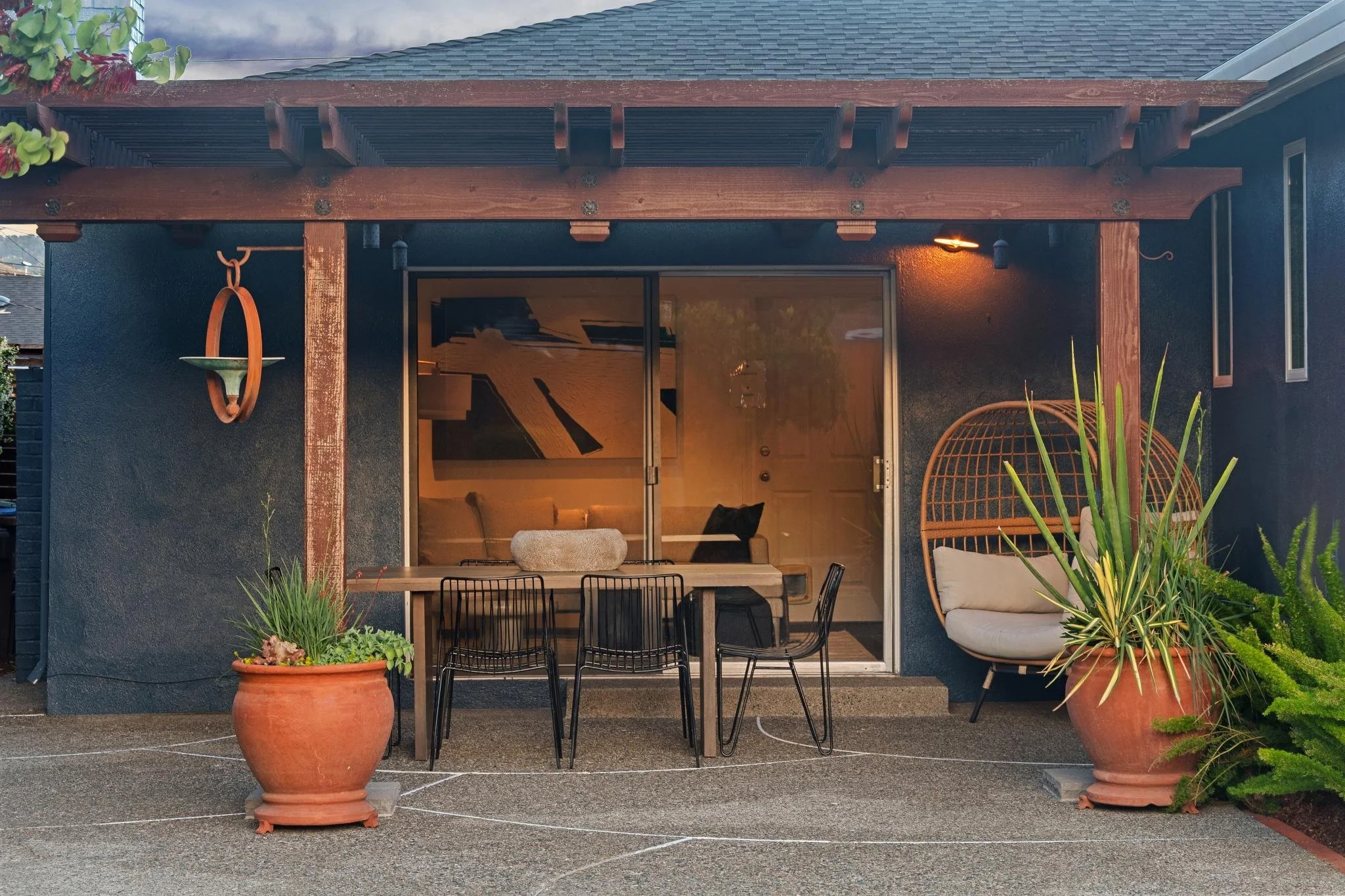A backyard patio with a covered pergola, outdoor dining table with chairs, and large potted plants including succulents. There is a sliding glass door leading into the house, and a cushioned patio chair on the right. The scene is in the evening with warm outdoor lighting.