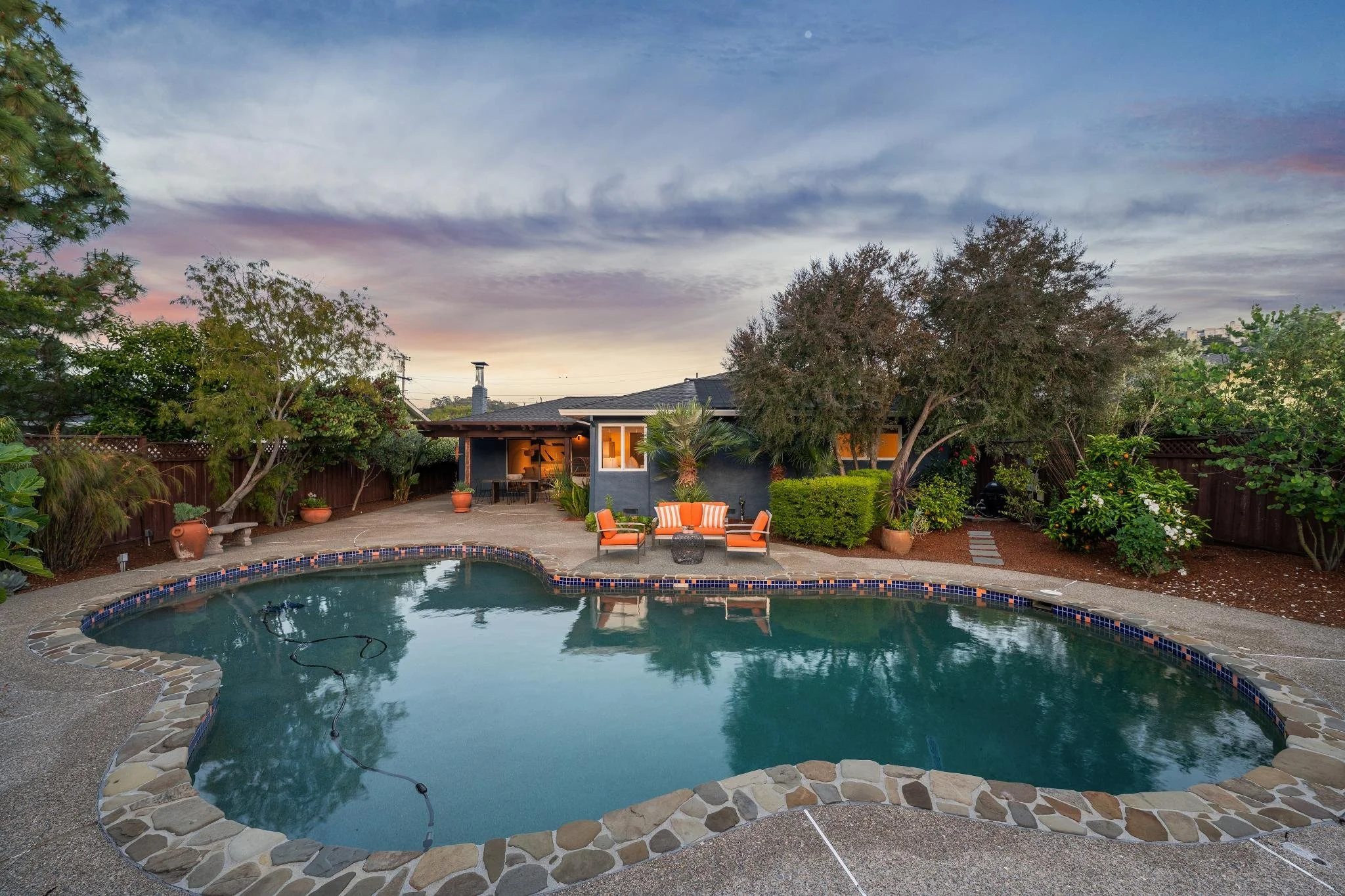 Backyard with swimming pool, patio furniture, and lush greenery at dusk with cloudy sky.