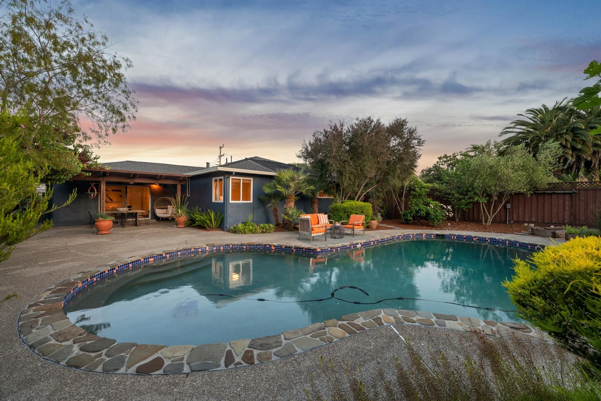 A backyard with a swimming pool, surrounded by trees and outdoor furniture, and a house with a patio, under a colorful sky at sunset.