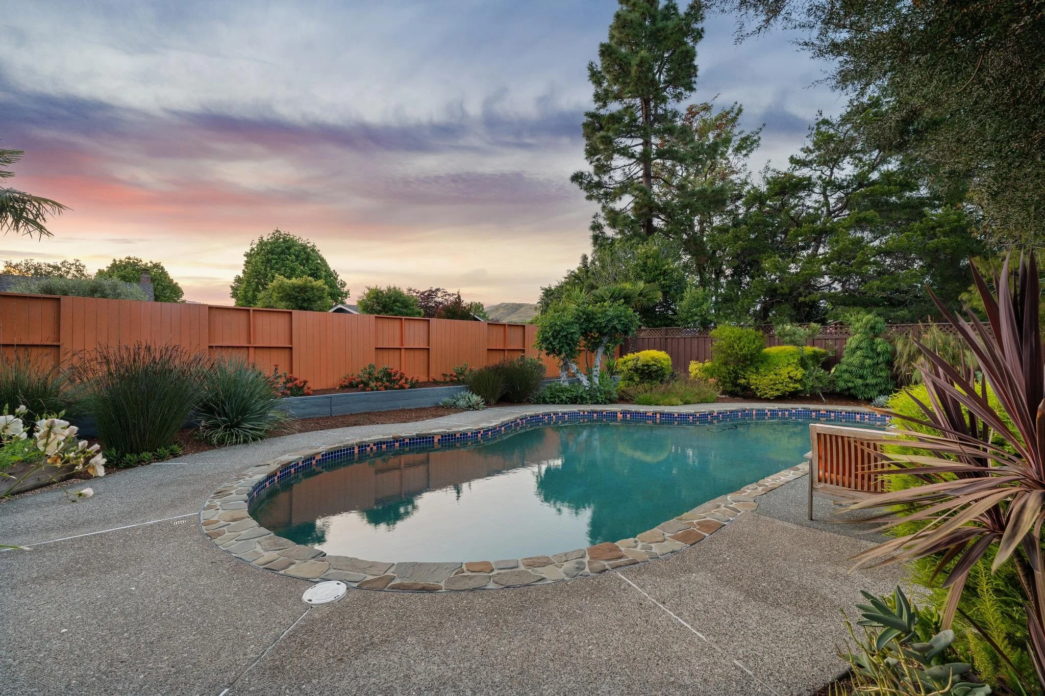 A backyard at sunset with a swimming pool surrounded by plants and trees, a wooden fence, and a scenic sky with clouds.