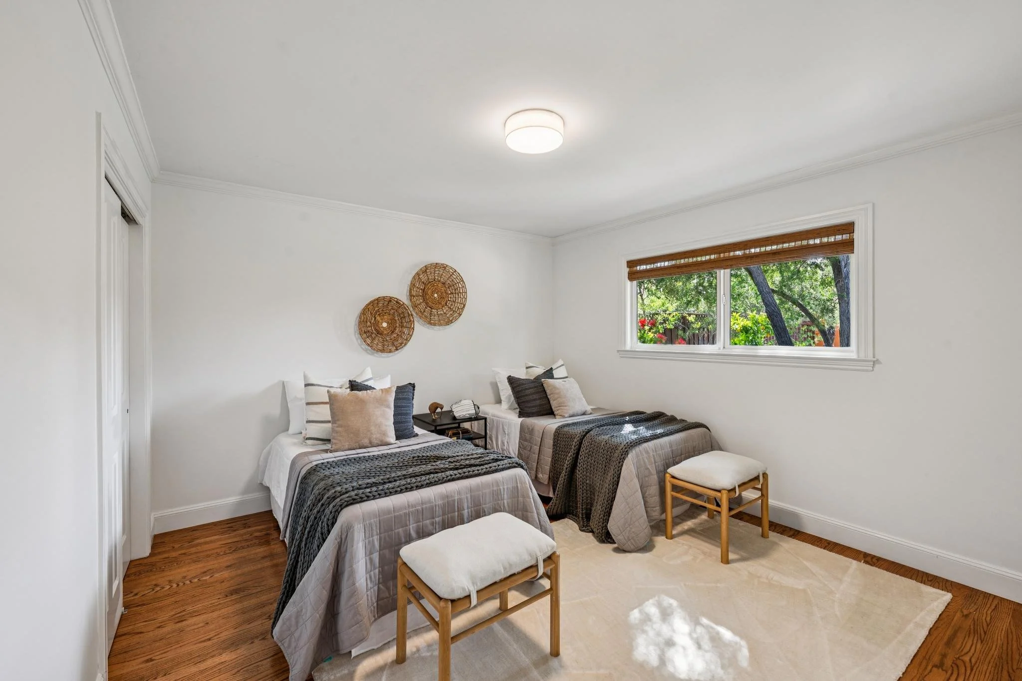 A bedroom with two twin beds, decorated with neutral-colored bedding and pillows, a small bedside table, and a window with brown blinds showing trees outside.
