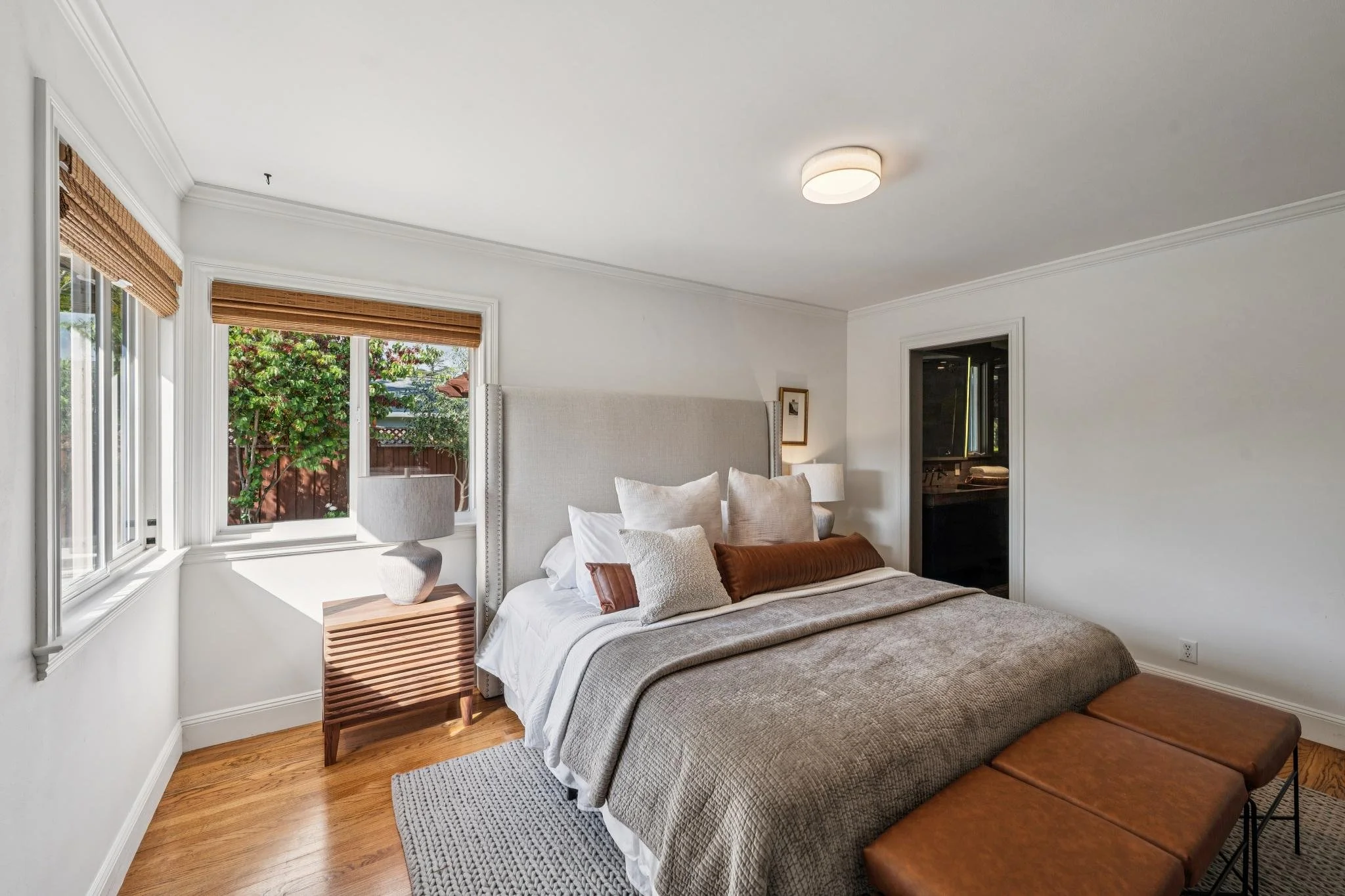 A bedroom with a white bed, tan and white pillows, wooden nightstand with a white lamp, window with bamboo shades, hardwood floor, white walls, and ceiling light.