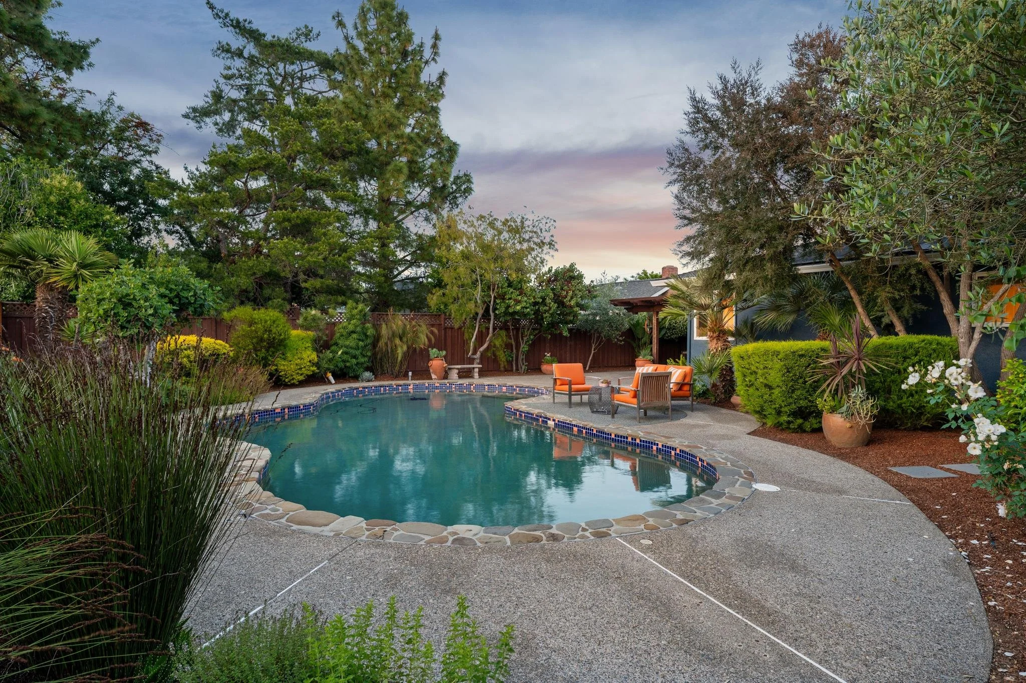 A backyard with a small, kidney-shaped swimming pool surrounded by a concrete patio. There are four orange chairs with a small table in between, and various potted plants. The yard is enclosed by a wooden fence, and there are tall trees and bushes al