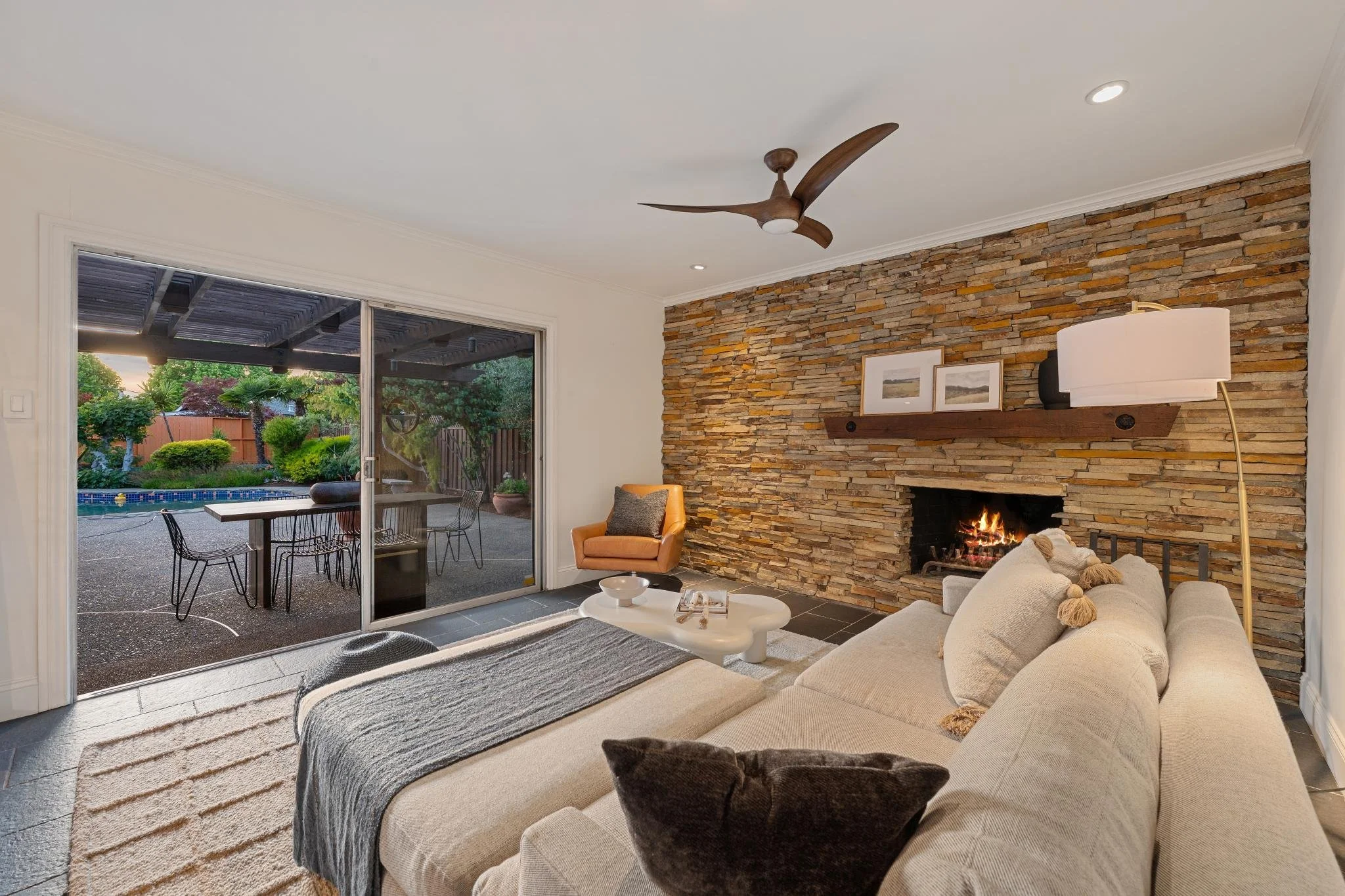 Living room with beige sofa, textured blanket, and decorative pillows, fireplace with stone wall, large sliding glass door opening to outdoor patio with garden and pool, ceiling fan, and modern decor.