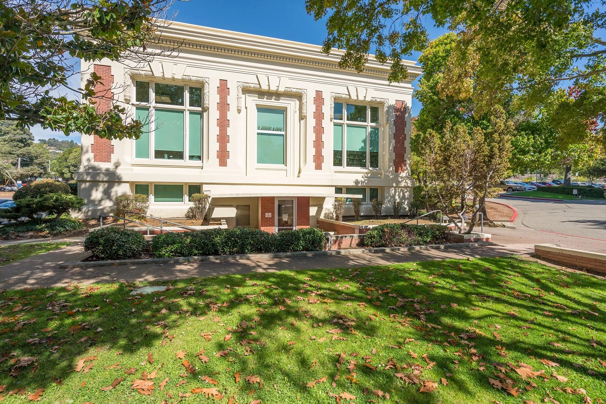The front of a historic white building with red brick accents, large windows, and decorative architectural details, surrounded by trees and a landscaped area with green grass and fallen leaves.