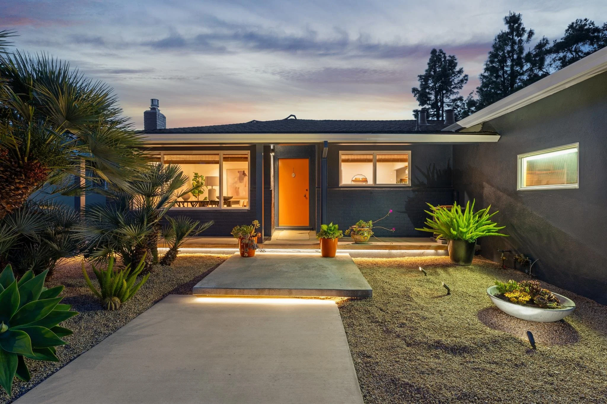 A house at dusk with illuminated interior, a concrete pathway leading to the front door, surrounded by desert plants, potted plants, and large windows.