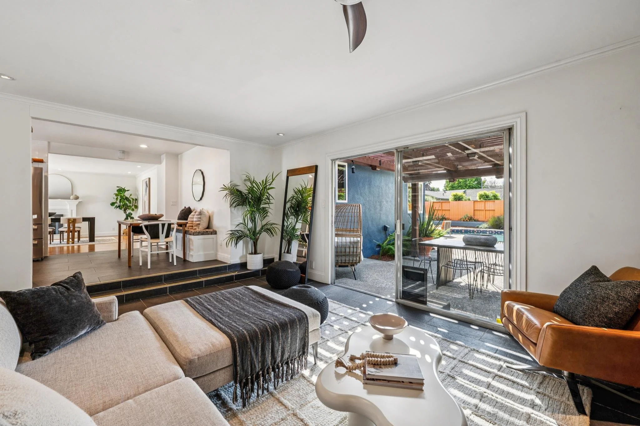Living room view showing a large beige sectional sofa with dark pillows, a white coffee table with books and decorative items, and a tan leather armchair. Large sliding glass doors lead to a backyard patio with outdoor furniture and a pool, with gree