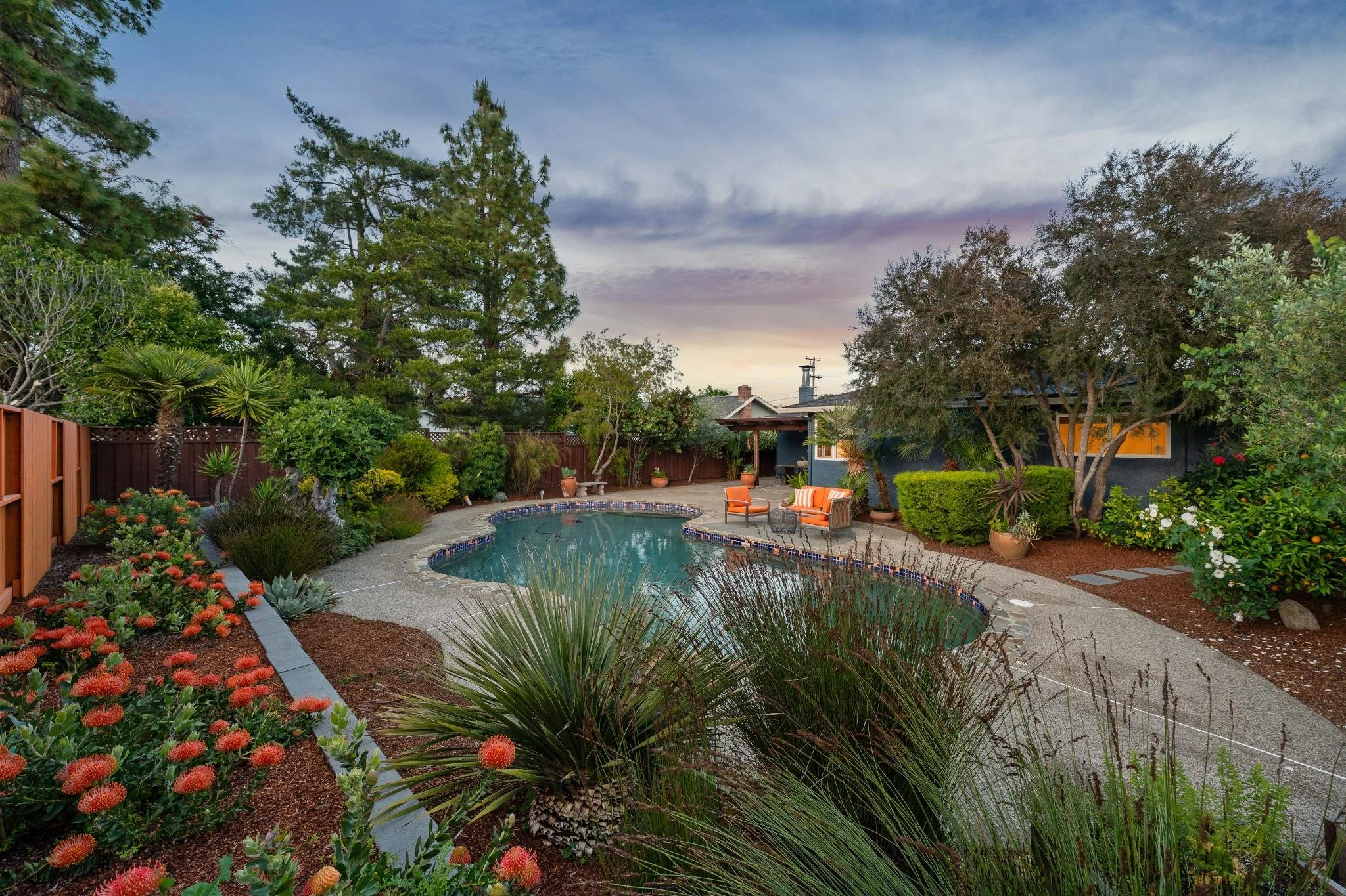 Backyard with a kidney-shaped swimming pool, surrounded by colorful flowers, shrubs, and trees under a cloudy sky.