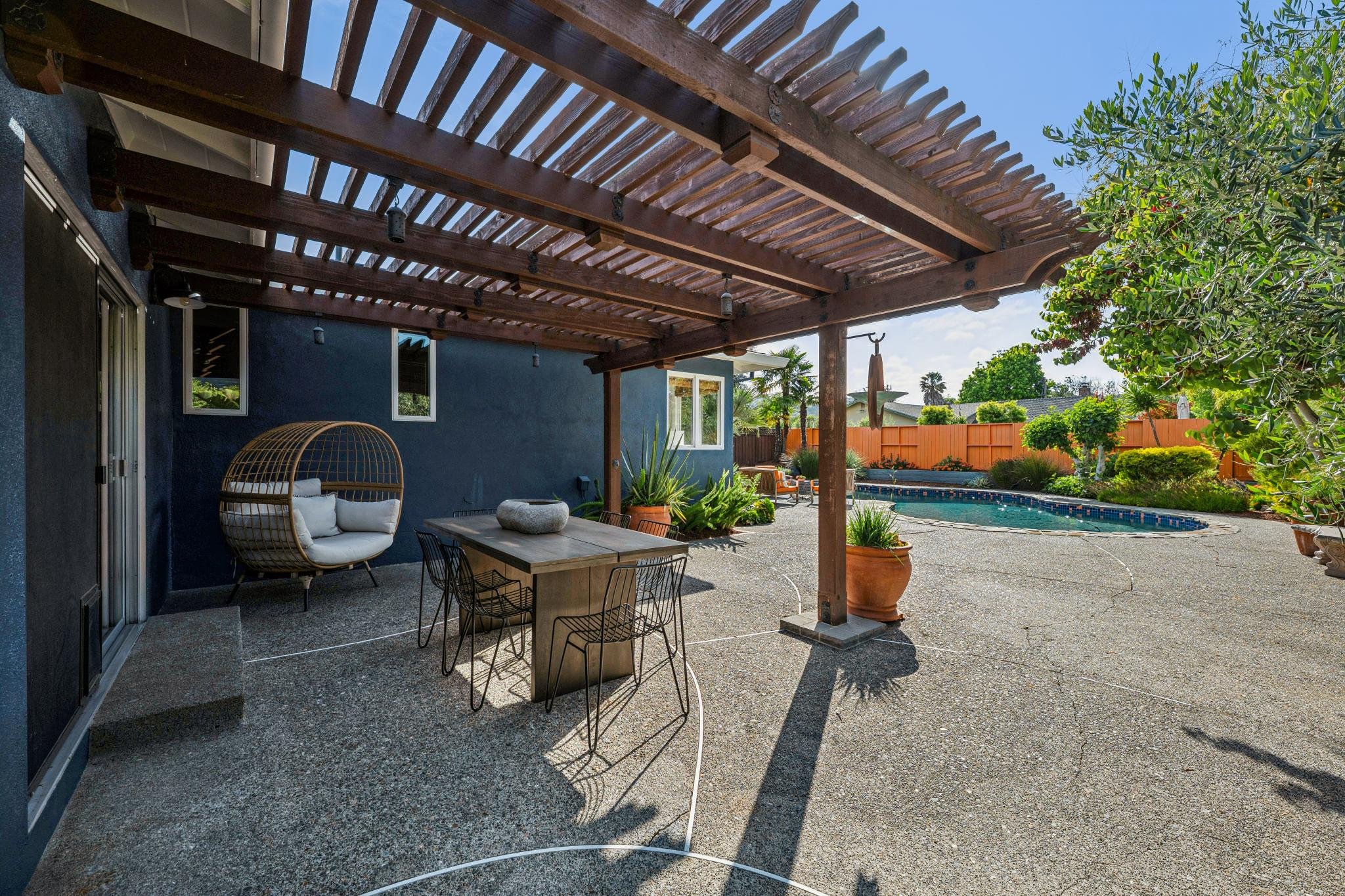 A backyard patio area with a wooden pergola, outdoor seating, dining table, and a swimming pool surrounded by plants and trees.