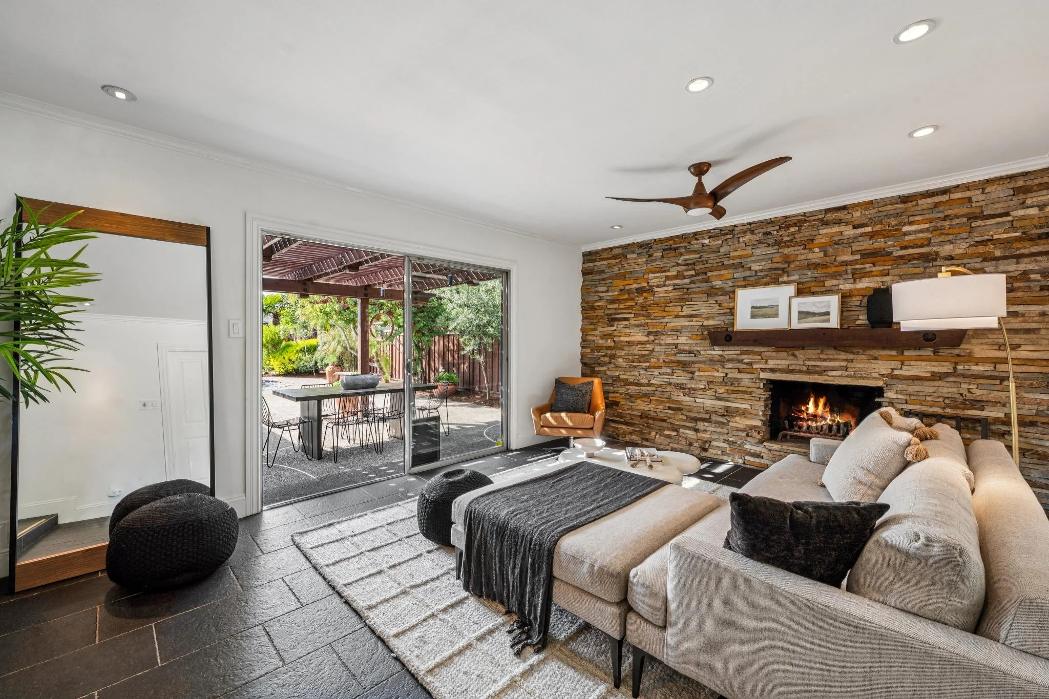 Living room with a stone fireplace, beige sofa with pillows, wooden ceiling fan, large sliding glass door leading to patio with outdoor furniture, potted plants, and garden view.