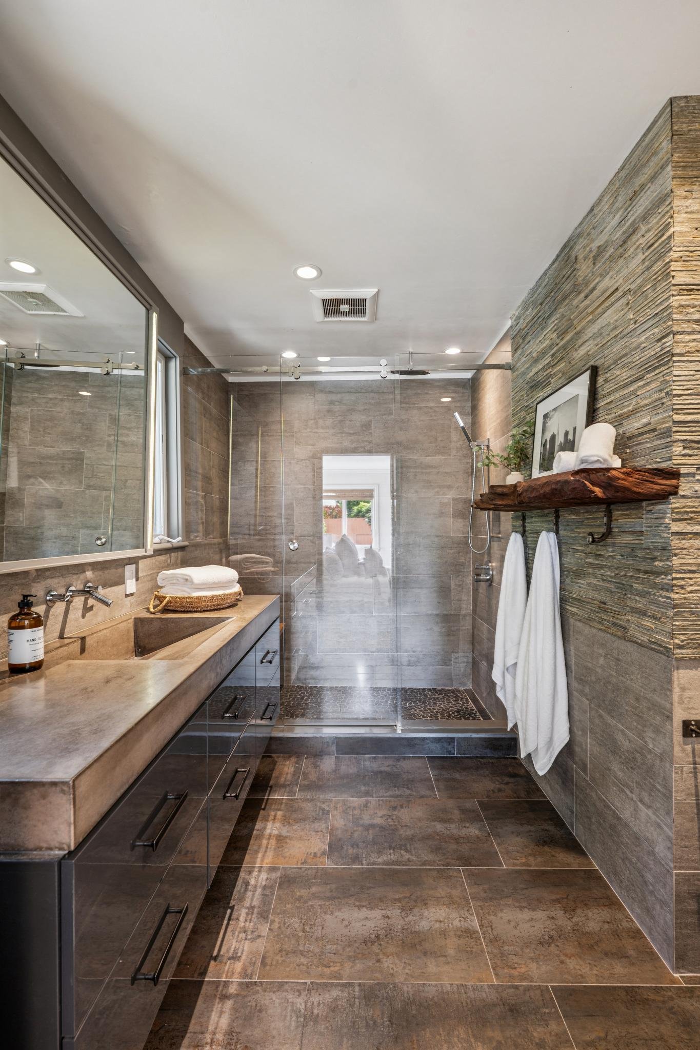 Modern bathroom with beige and brown tiled walls and floors, a glass shower enclosure at the end, a vanity with a beige countertop and black cabinets, and a wooden shelf with decorative items and towels.
