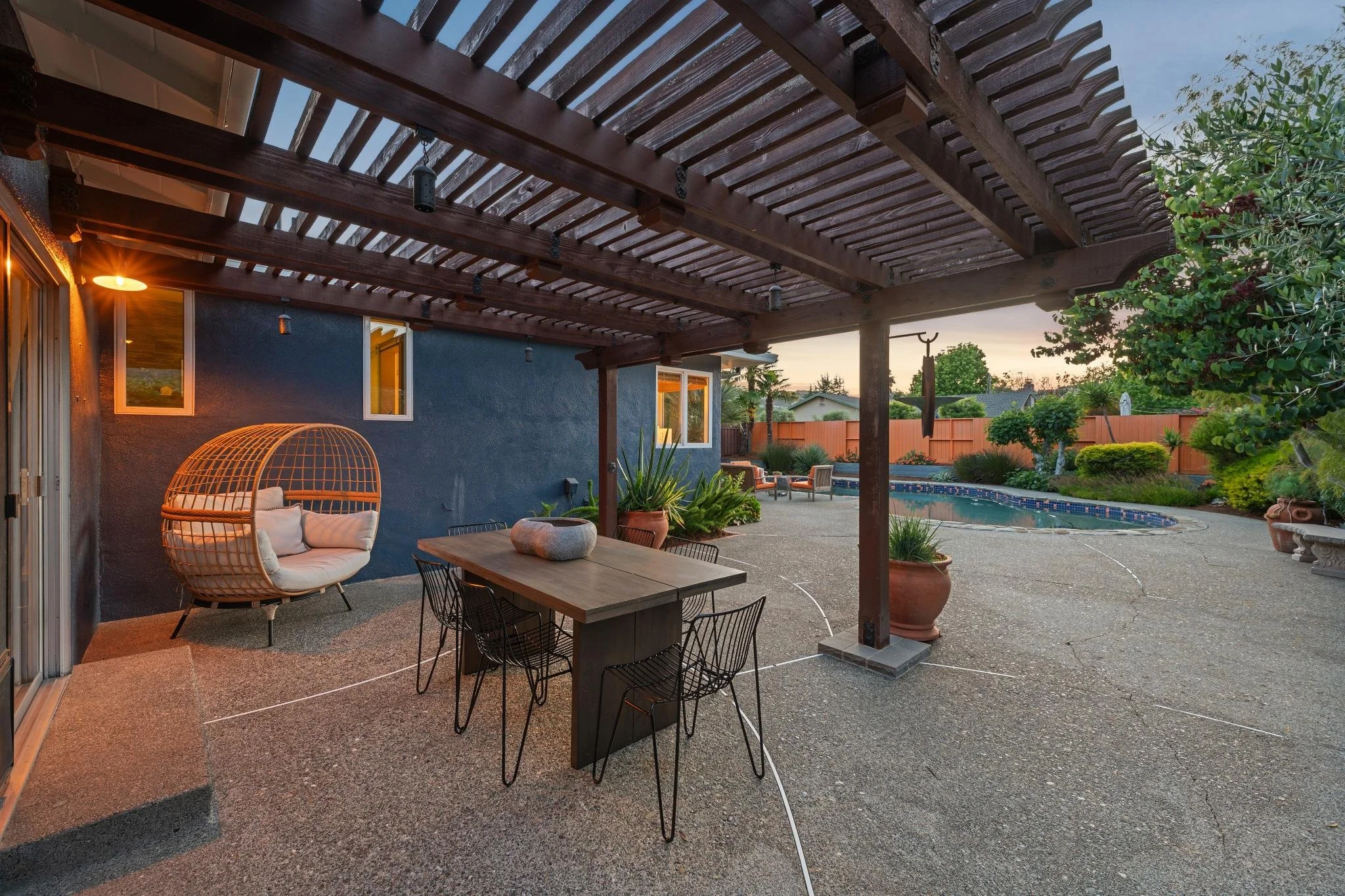 A backyard patio area with a covered wooden pergola, outdoor furniture including a cushioned papasan chair and a dining table with chairs, potted plants, a swimming pool, and a landscaped yard with trees and shrubs at sunset.