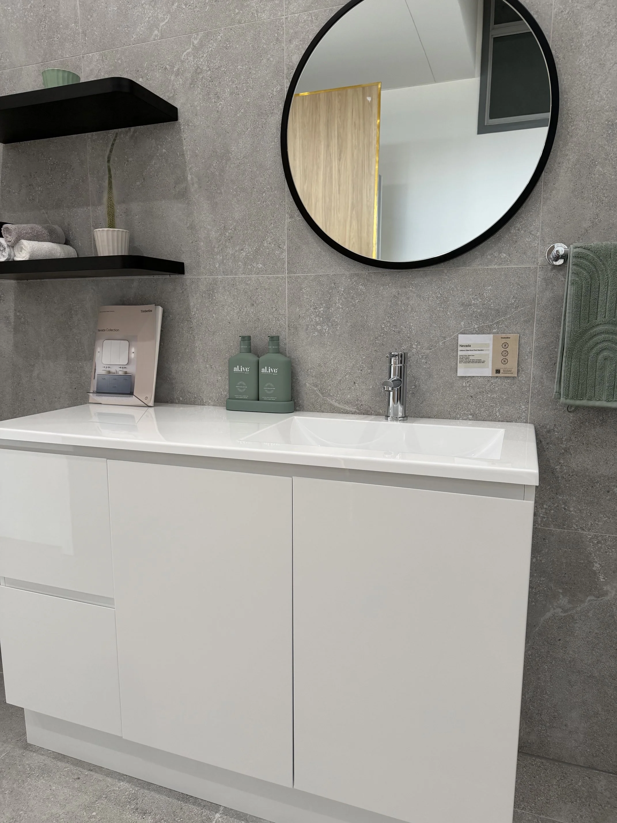 Modern bathroom with a white vanity, a round mirror, grey tiled wall, and two green bottles of skincare products.