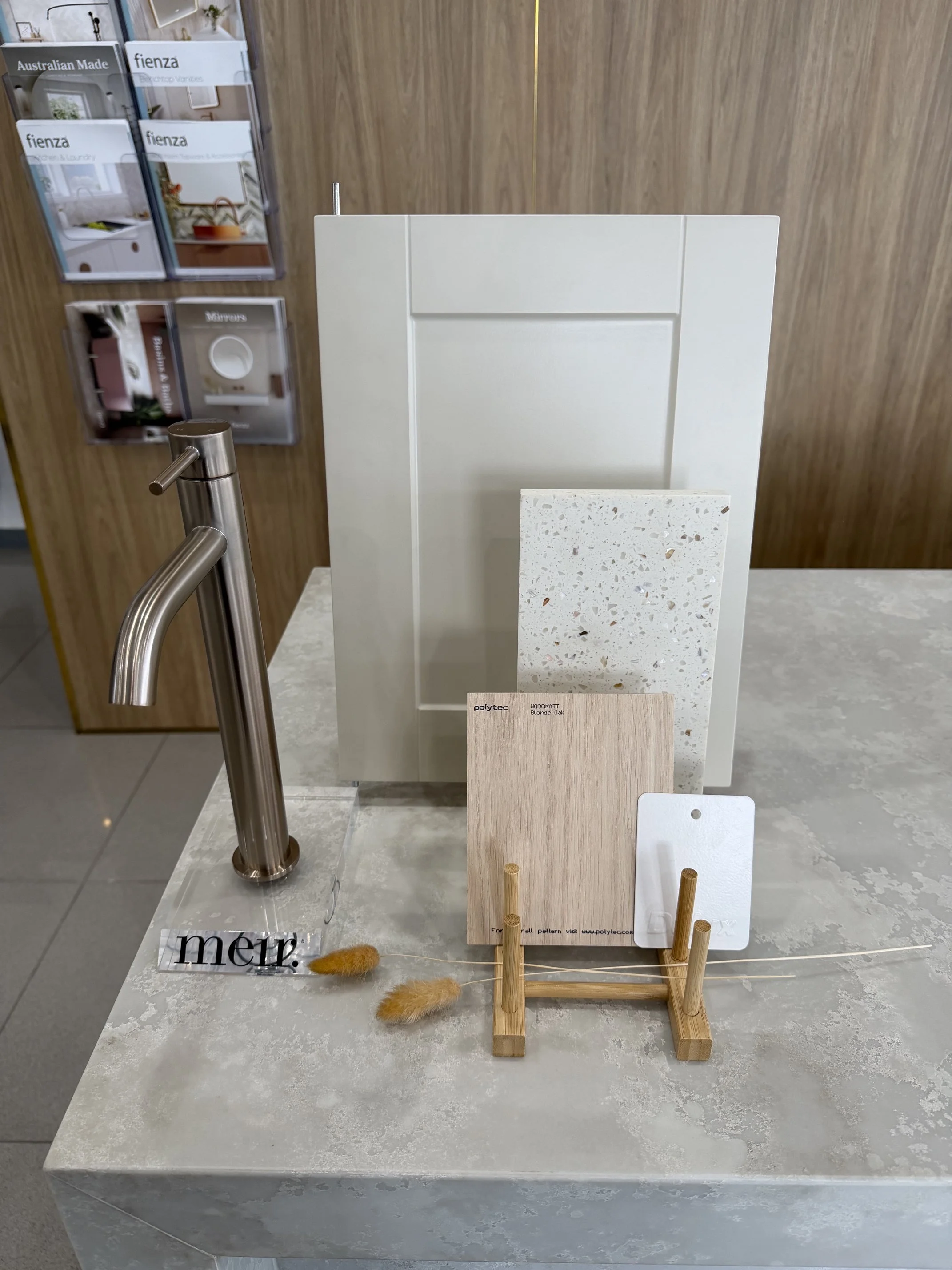 Display of various interior design material samples on a grey marble table, including tiles, wood, and aggregate finishes. Magazines and a metal faucet are visible in the background.