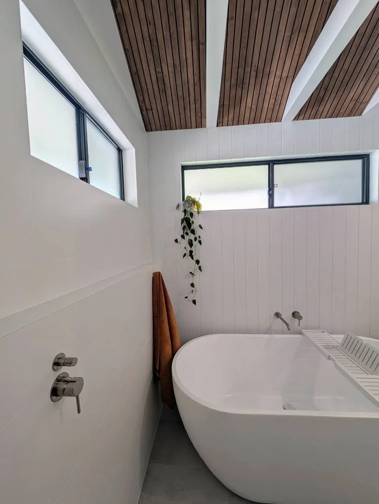Modern bathroom with a white bathtub, a brown towel hanging on the wall, a small potted plant, and three narrow horizontal windows, with a wooden ceiling and white wall paneling.