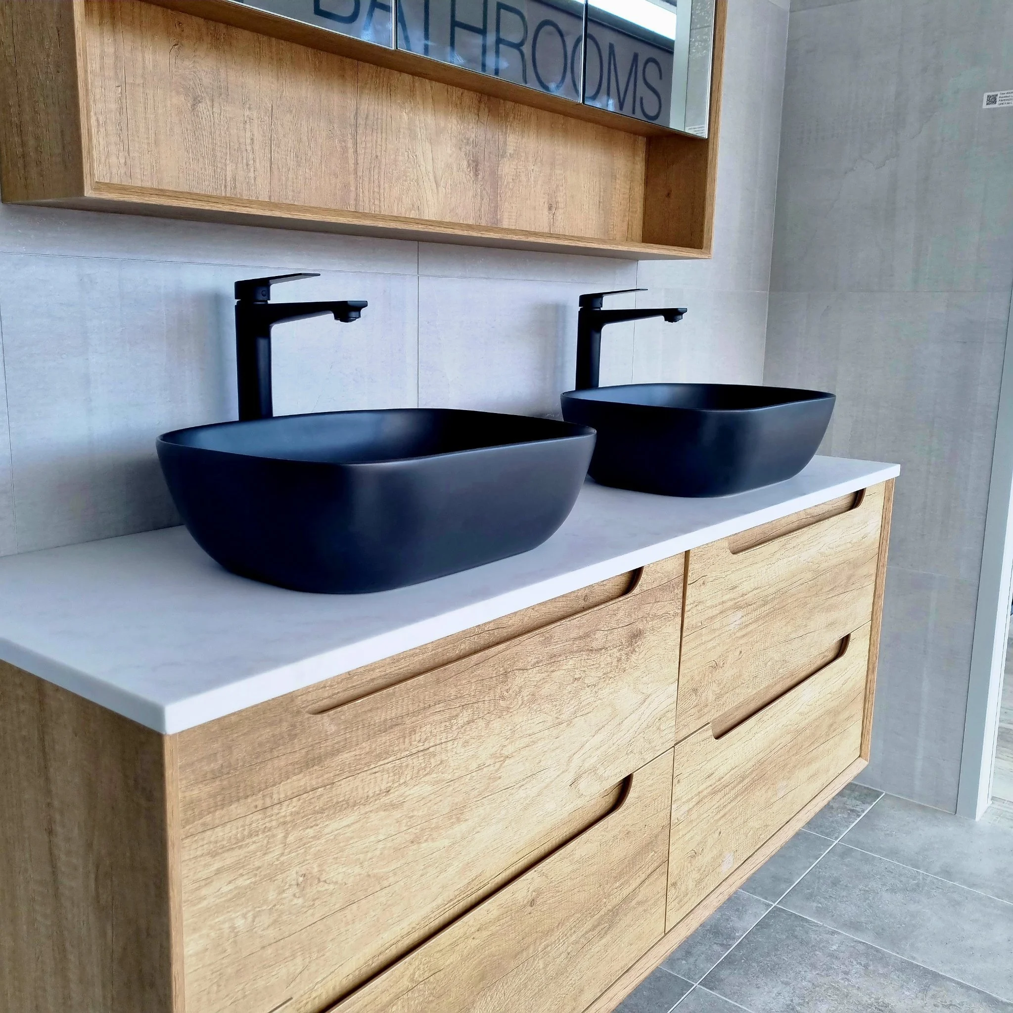 Modern bathroom with two black vessel sinks, black faucets, and a wooden cabinet with a white countertop. The wall behind the sinks has a mirror and a QR code sticker, with tiled flooring.