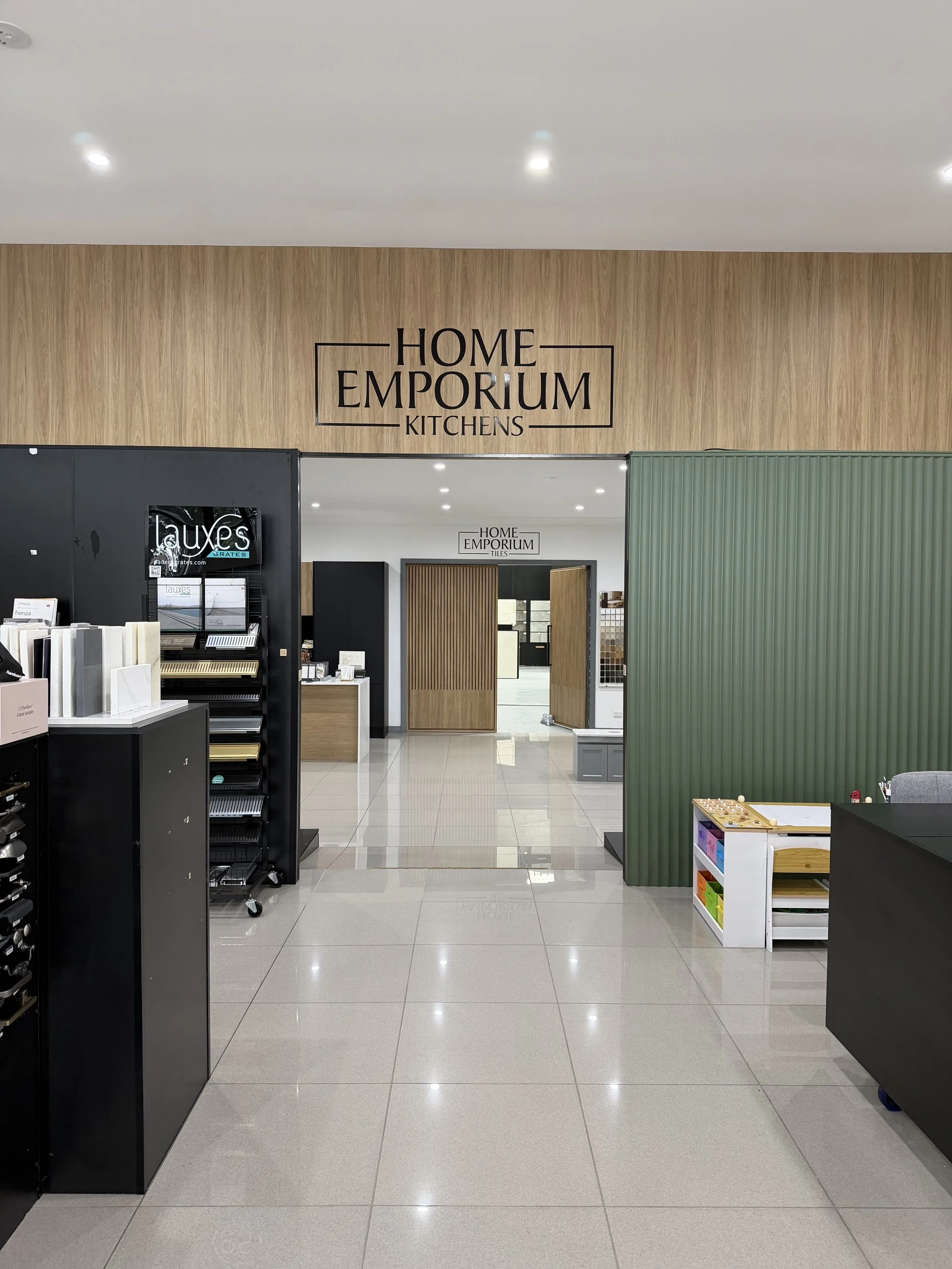 A home improvement store interior with a sign reading 'Home Emporium Kitchens'. Display shelves with color samples, a green partition, and a doorway to another section.