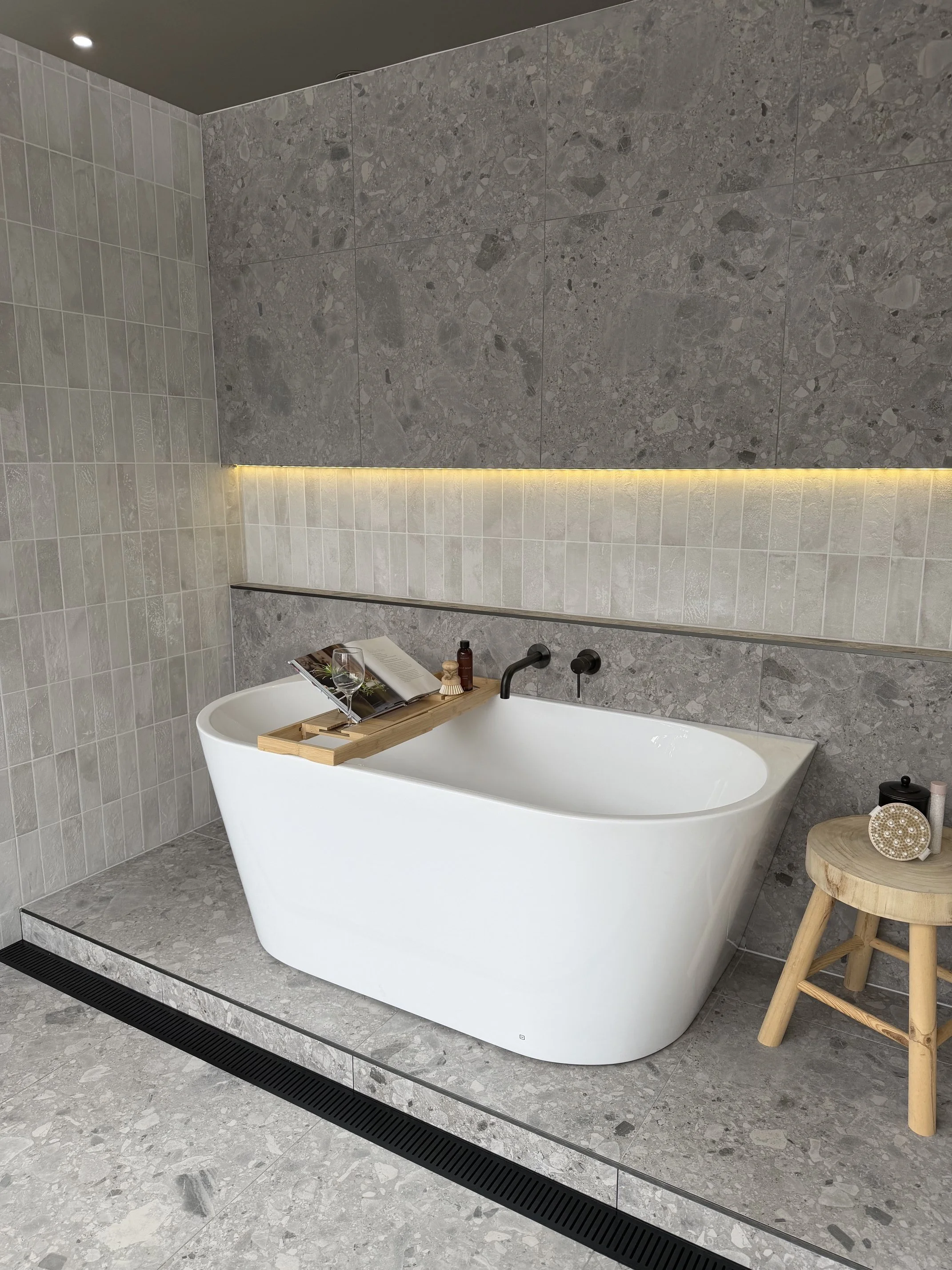 Modern bathroom with a white bathtub, gray stone wall and floor, a small wooden stool with toiletries, and a wooden bath tray with a glass, magazine, and bottles. There is a black drainage grate on the floor and warm lighting above the tub.