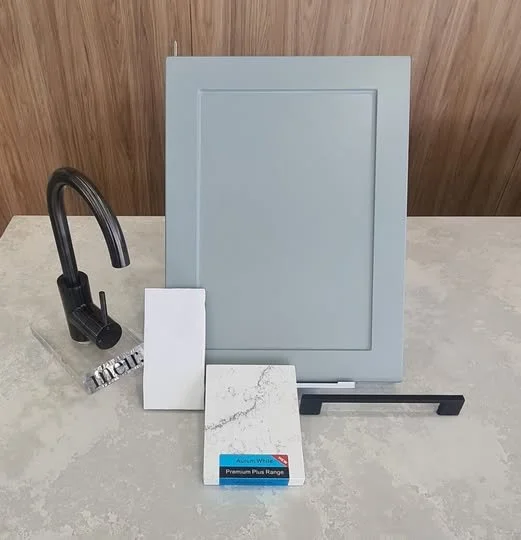 A sample display with a black kitchen faucet, three sample tiles, a grey framed panel with a stand, and a black sample stand on a light countertop.