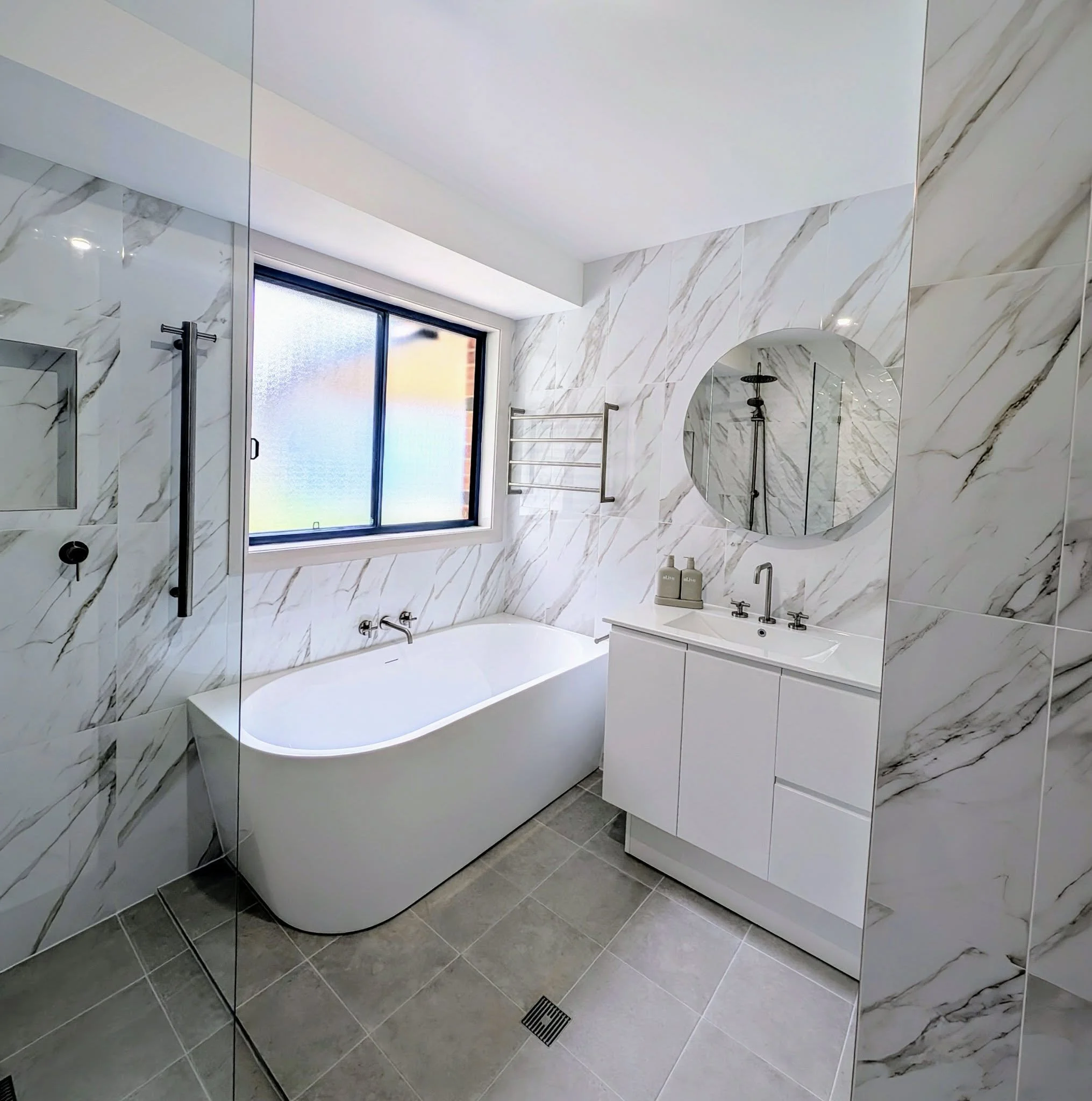 Modern bathroom with marble walls, a large mirror, a window, and a bathtub. There is a towel rack and a sink with a countertop. The bathroom has gray floor tiles and polished surfaces.